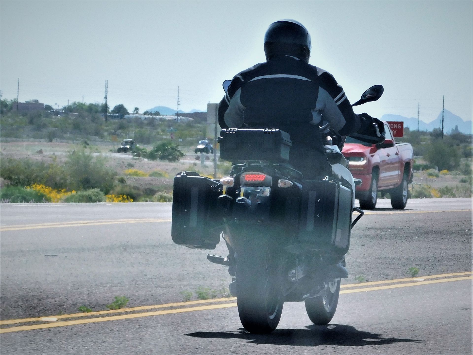 Motorcyclist on a road, back view, wearing black gear, passing a red truck on a sunny day.