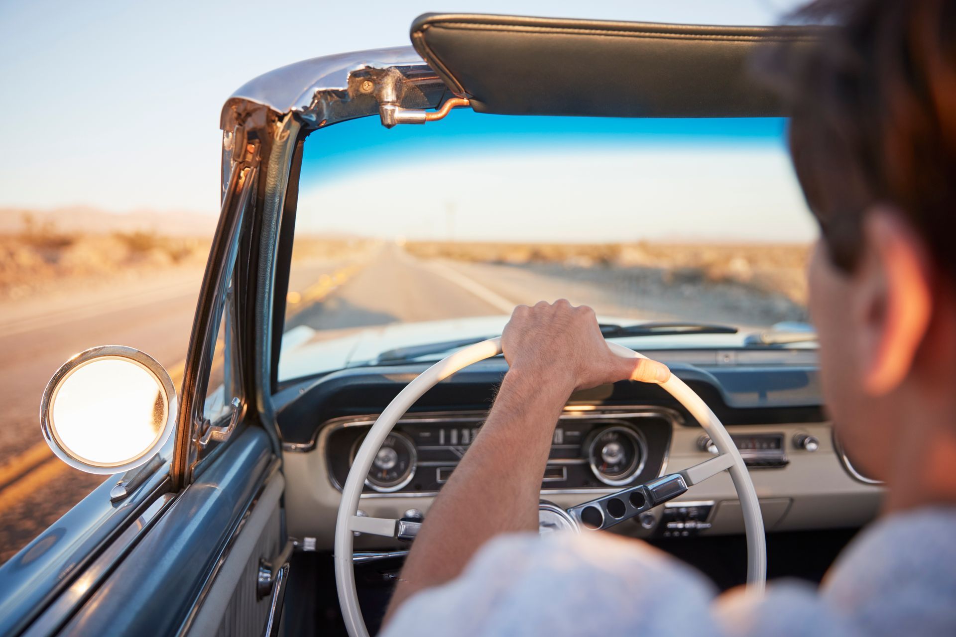 Person driving a convertible down a desert road, with a blue sky overhead.