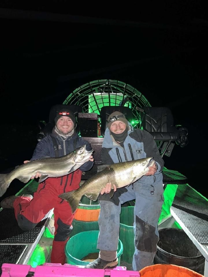 Two men are holding fish on a boat at night.