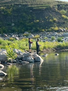 Two people are fishing in a river near a rocky shoreline.