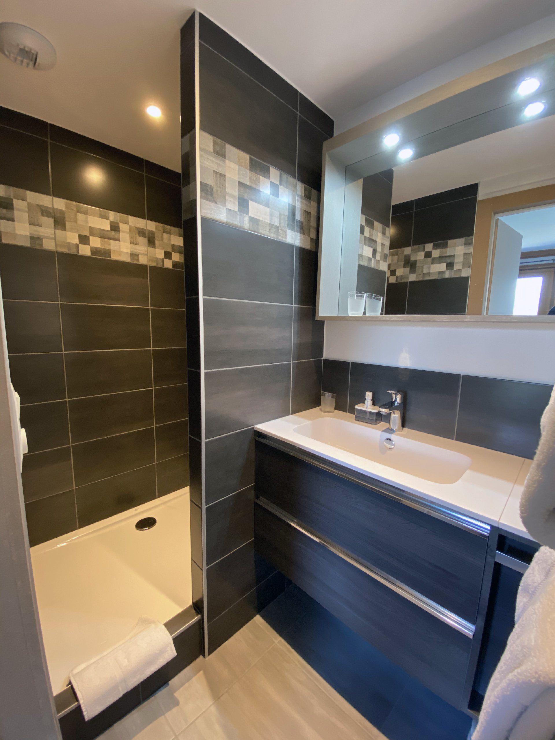 Salle de bains moderne avec douche carrelée noire, baignoire blanche et meuble-lavabo foncé avec miroir.