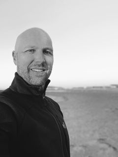 Un homme chauve souriant, vêtu d'un pull foncé, se tient à l'extérieur, au bord d'une plage, par une journée nuageuse.