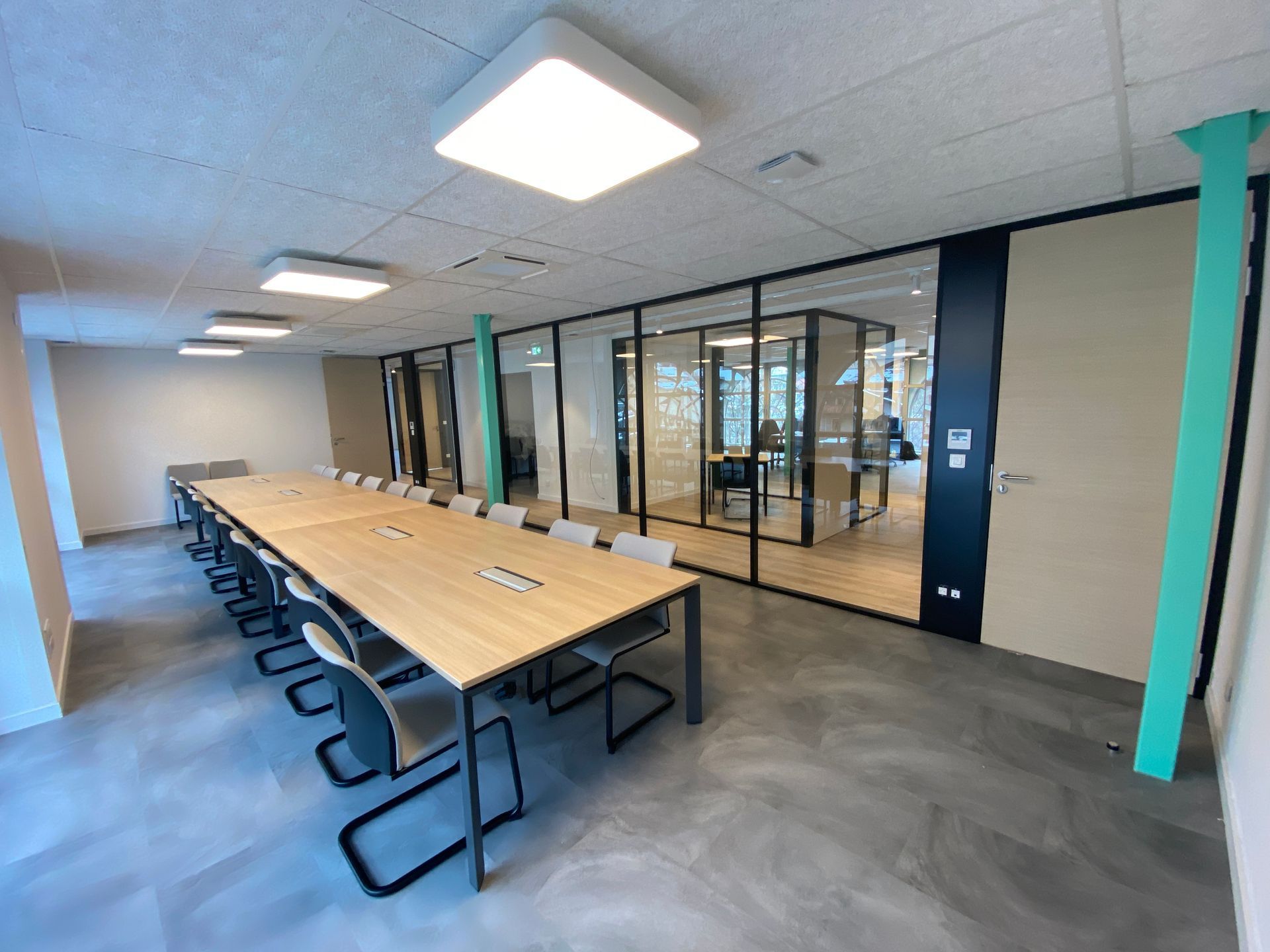 Salle de réunion moderne avec une longue table en bois, des chaises noires, des parois vitrées et des colonnes turquoise.