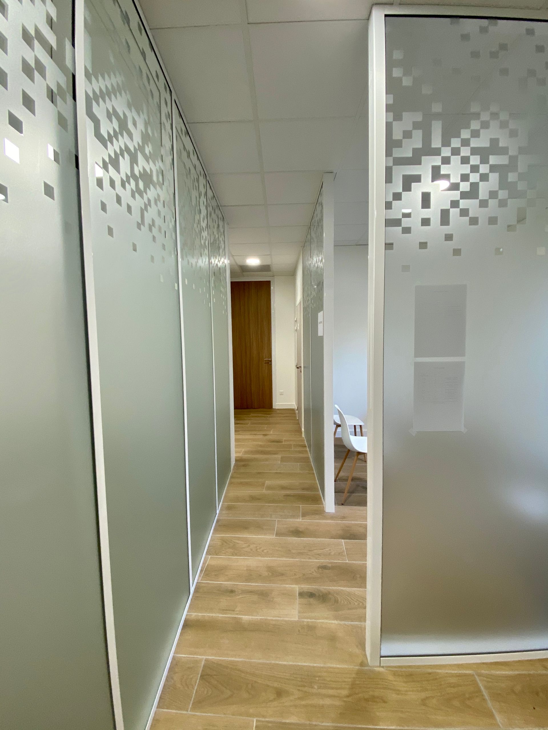 Couloir de bureau moderne avec parois en verre dépoli et sol imitation bois, menant à une porte en bois.