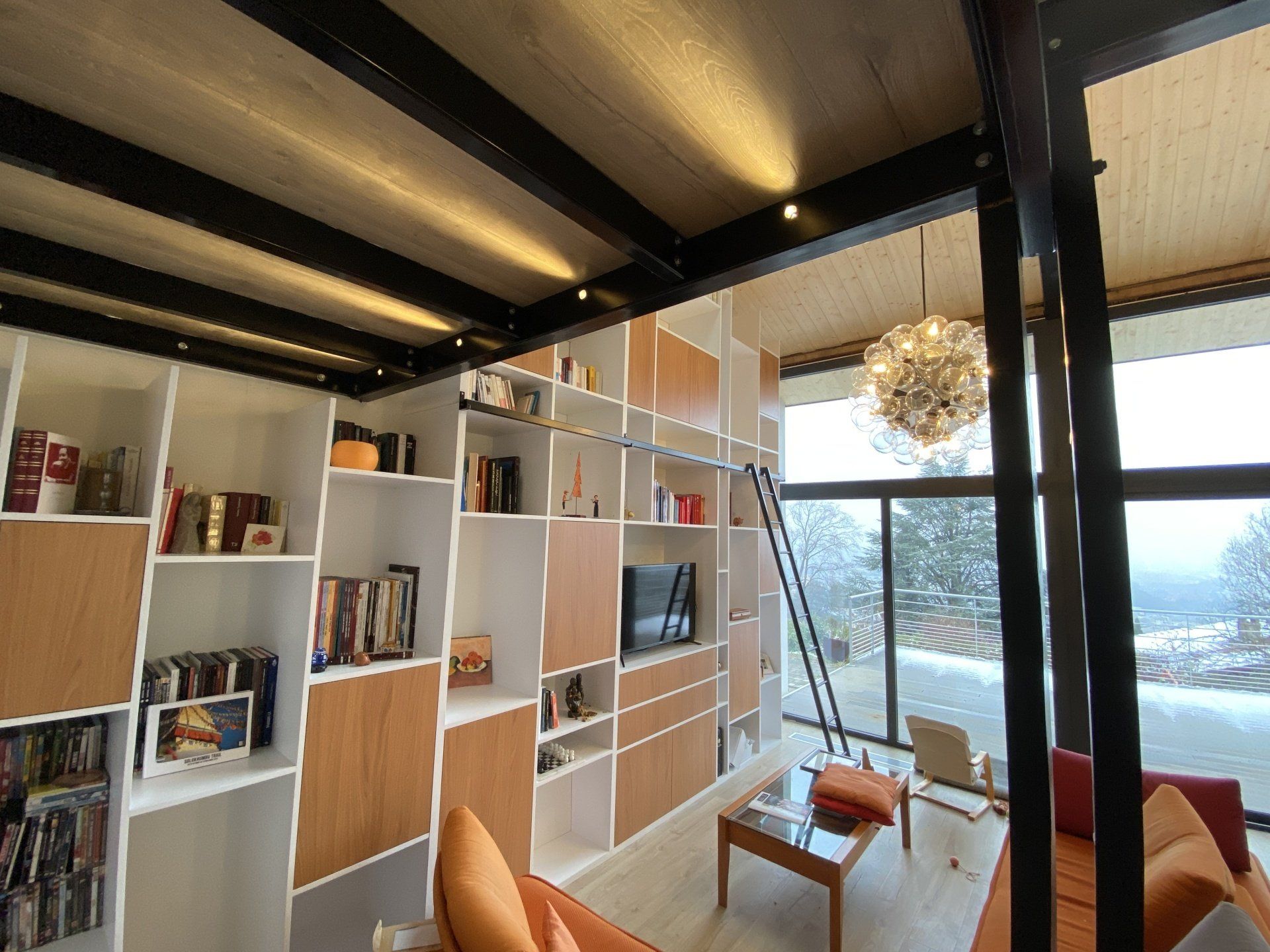 Salon loft moderne avec étagères blanches, chaises orange et grandes fenêtres donnant sur une vue