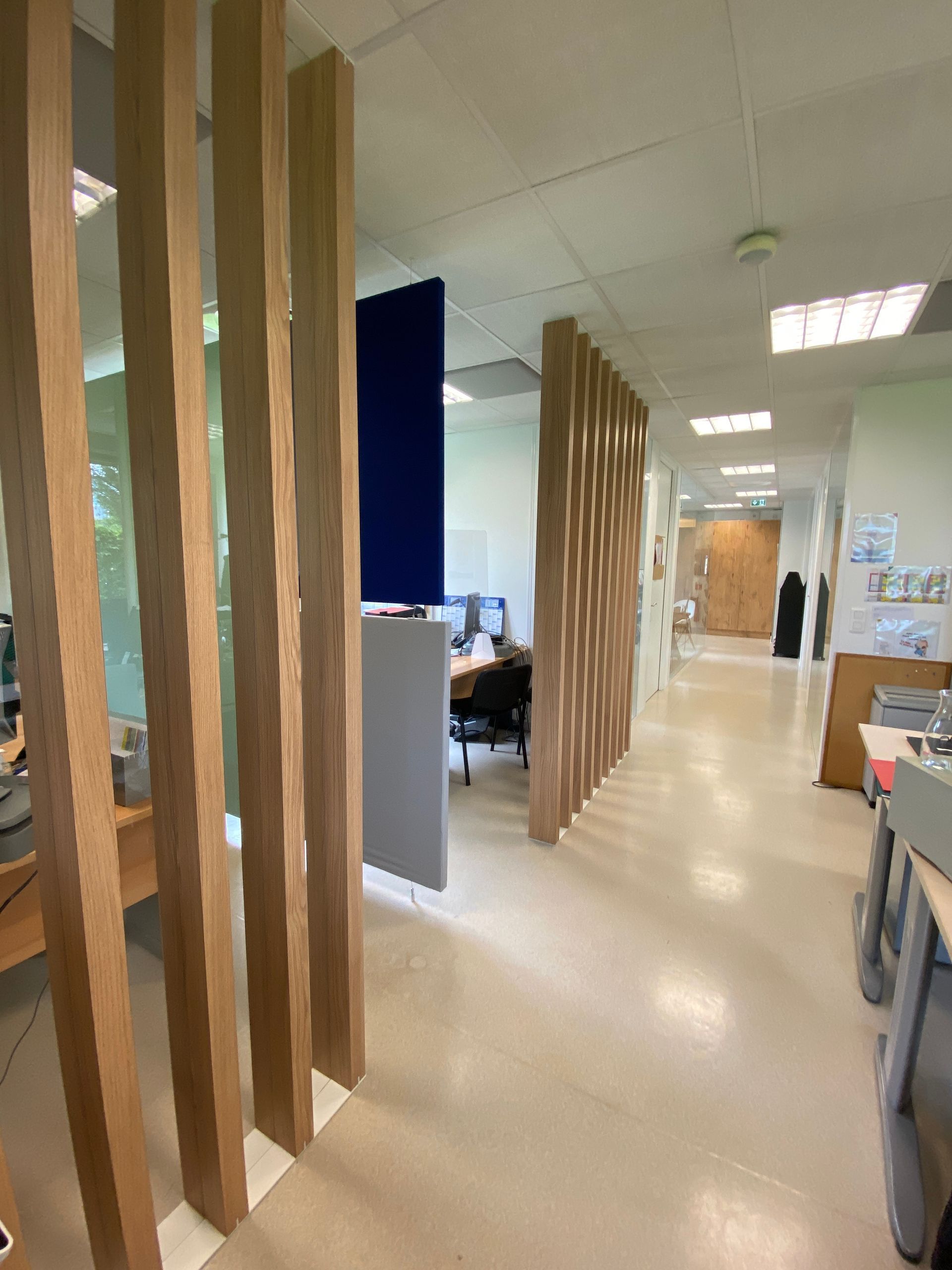 Couloir de bureau avec cloisons en lattes de bois, sol beige et bureaux éclairés par des plafonniers lumineux.