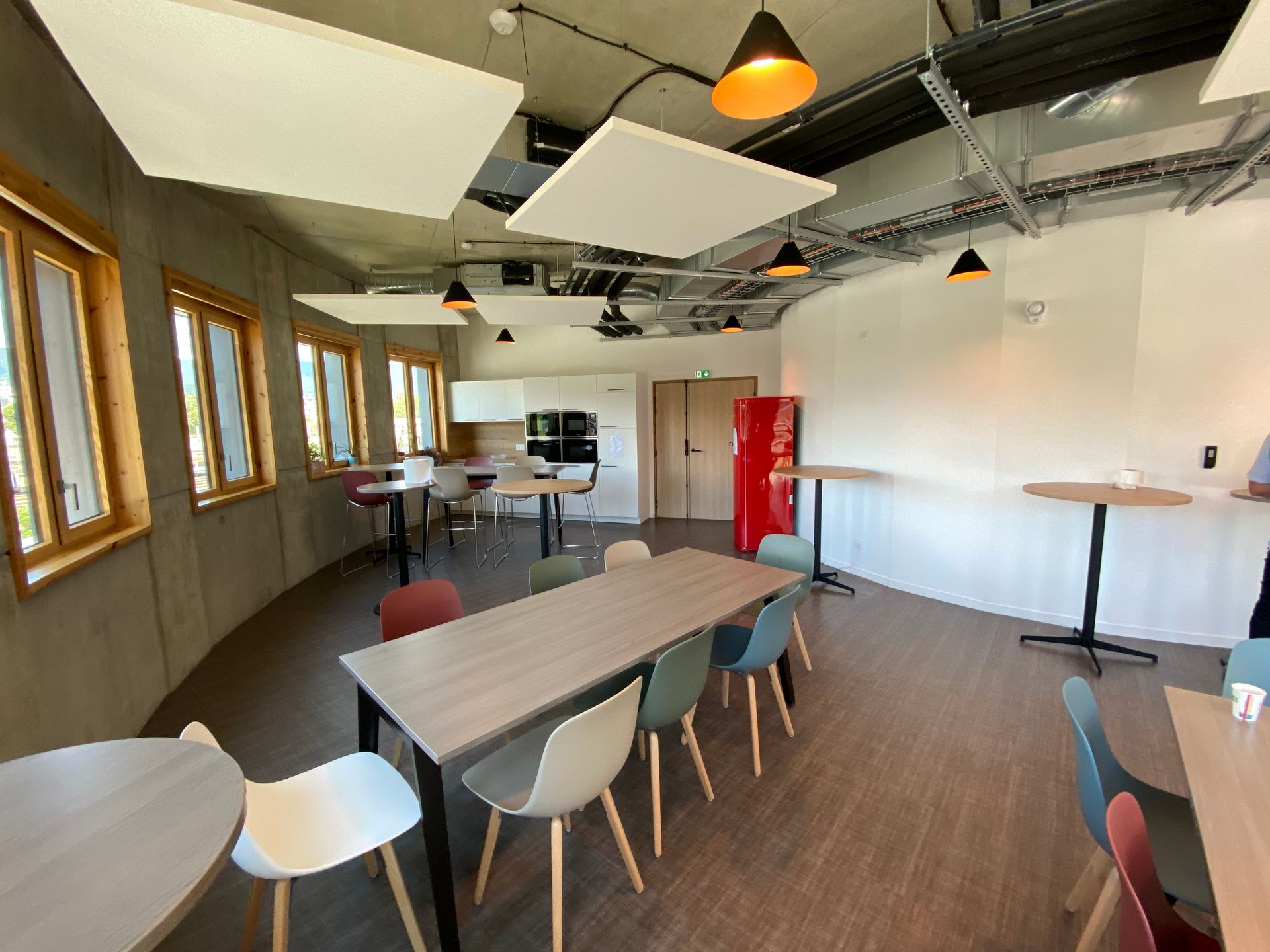 Salon de bureau moderne avec tables, chaises colorées, fenêtres et porte rouge sous des suspensions lumineuses