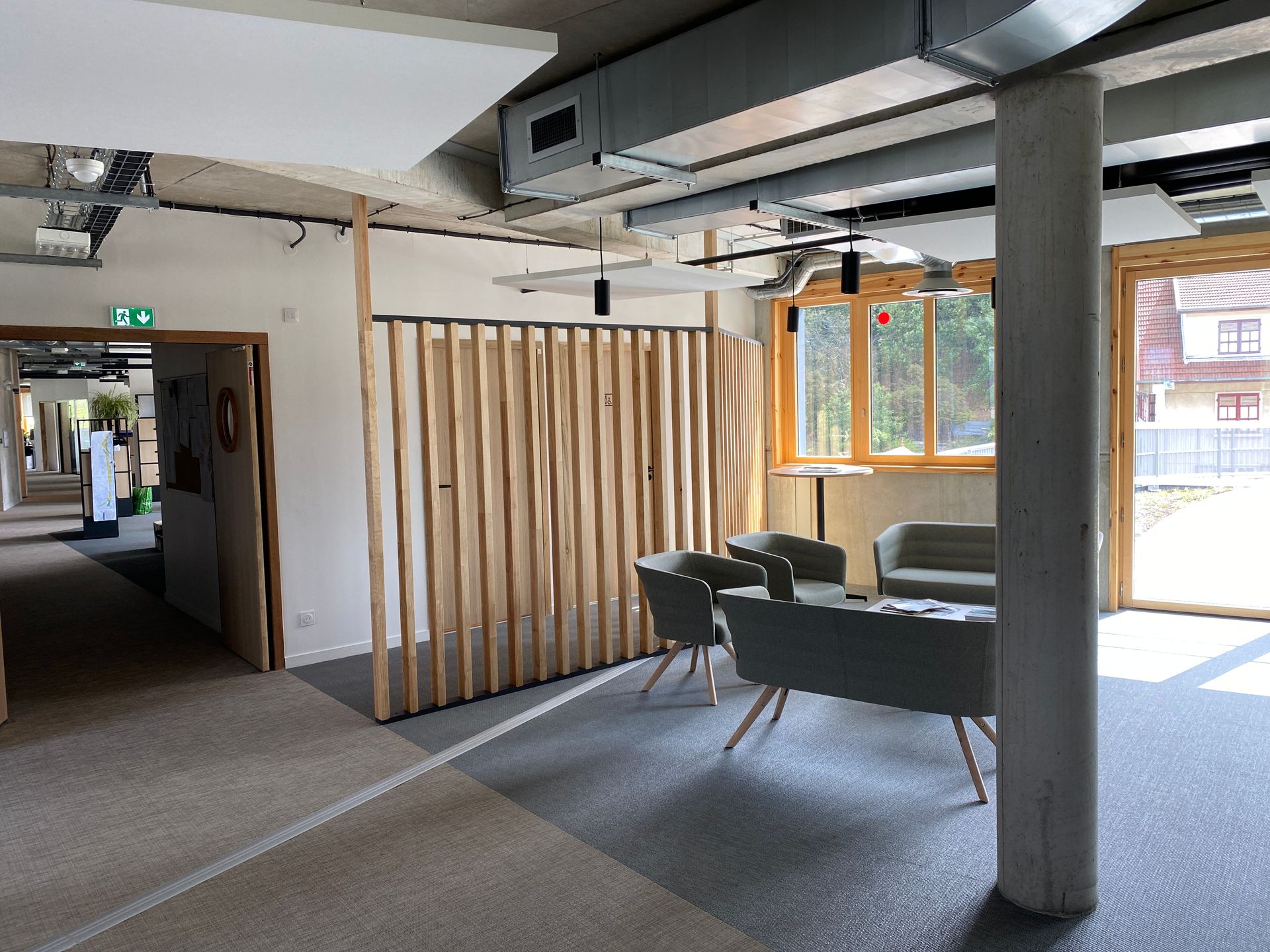 Espace lounge moderne avec canapés gris, fauteuils, cloison en bois et grandes fenêtres dans un espace de bureau lumineux