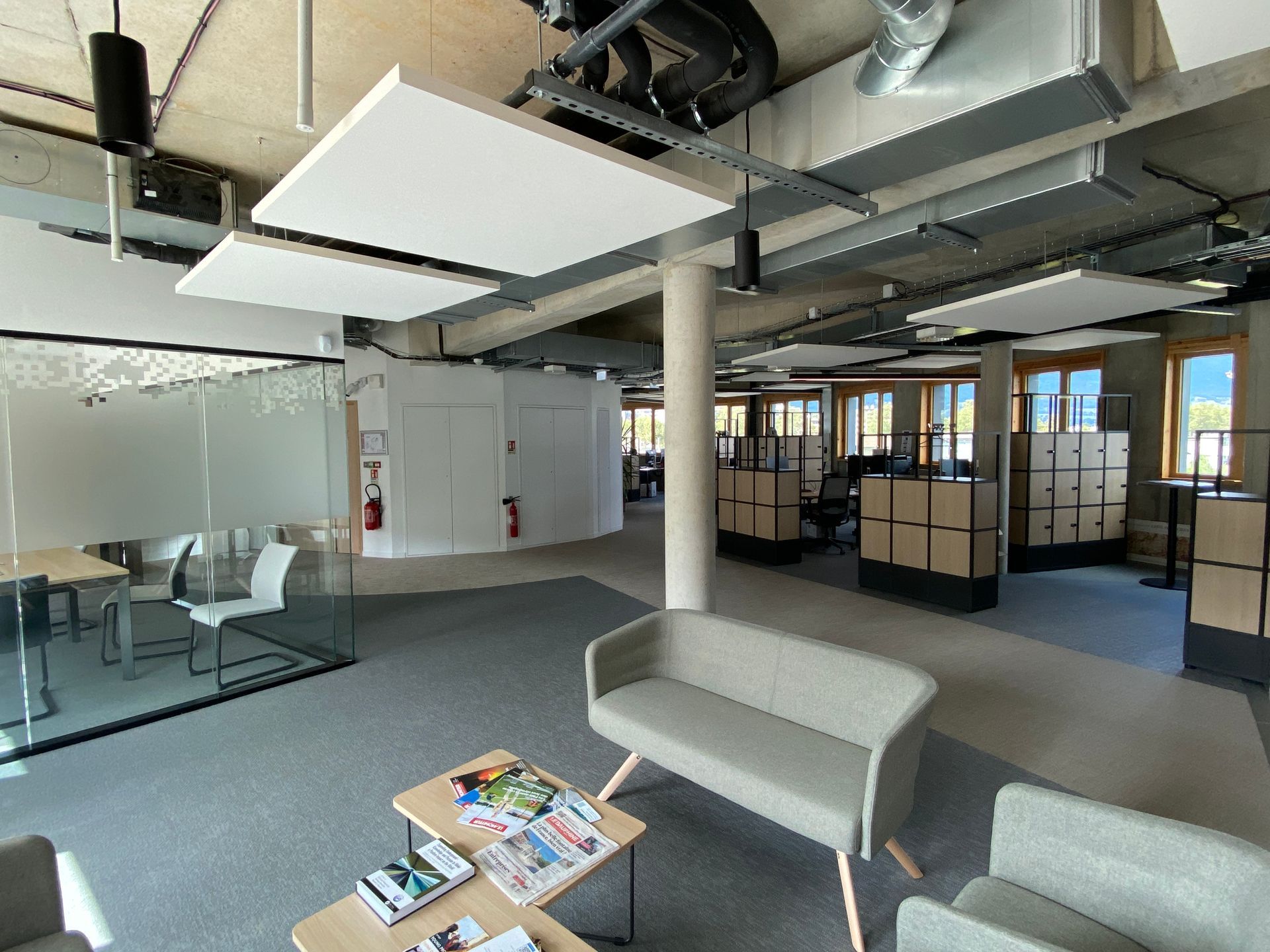 Salon de bureau moderne avec chaises grises, canapés, table basse, salle de réunion vitrée et plafond apparent.
