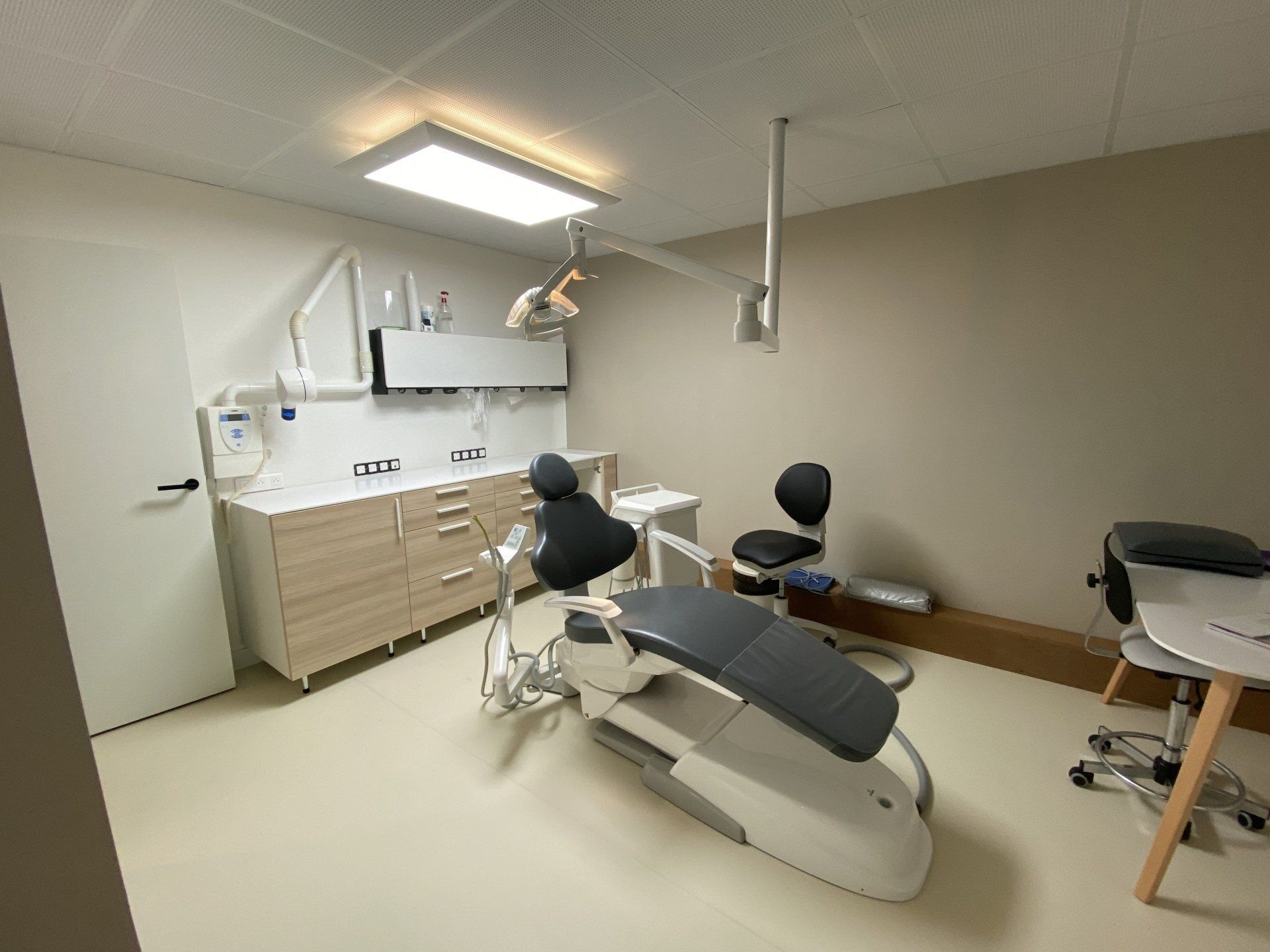 Salle d'examen dentaire avec fauteuil, éclairage au plafond, armoires et lavabo sur sol beige.