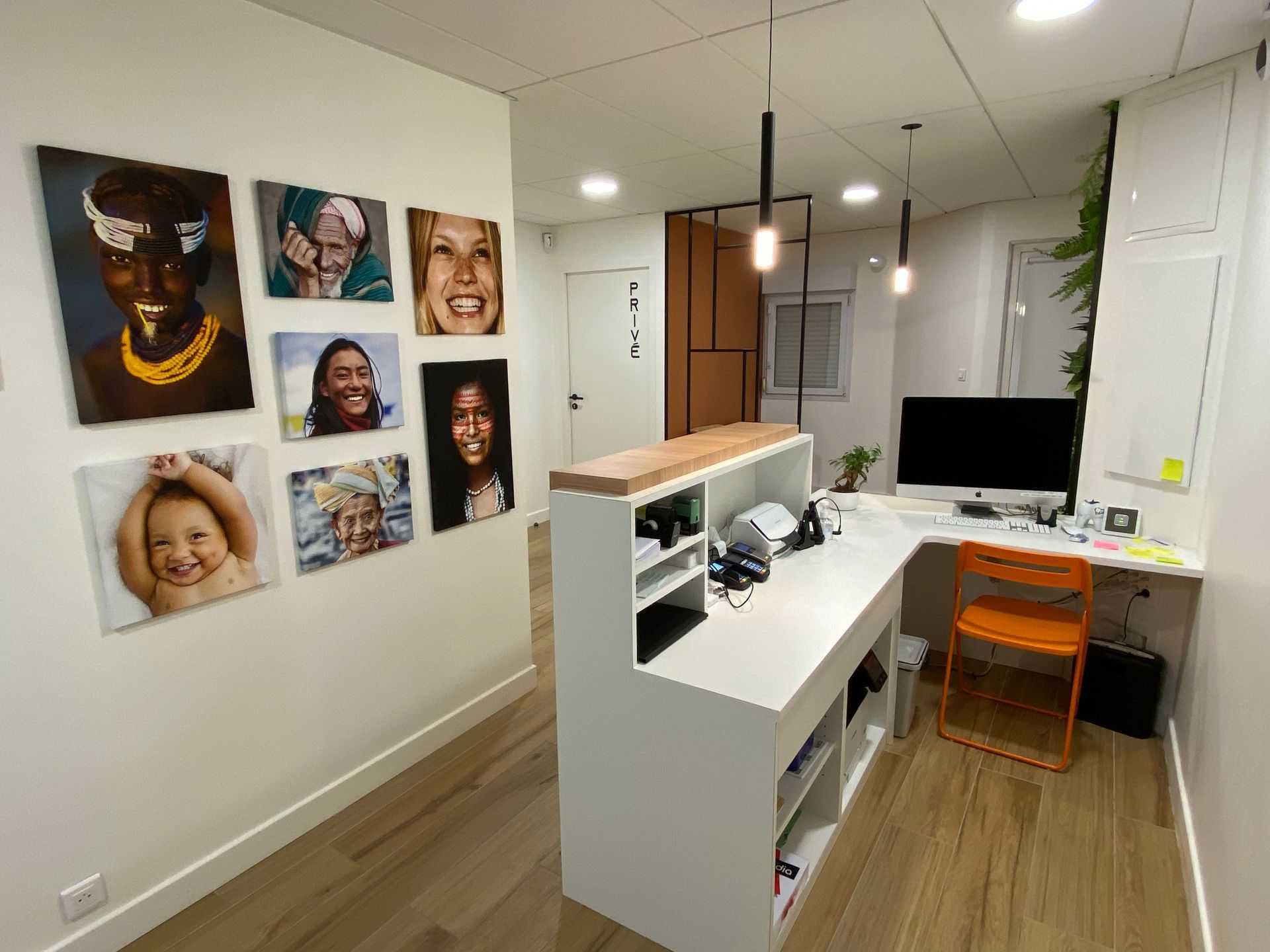 Espace d'accueil moderne avec bureau blanc, ordinateur, chaise orange et portraits encadrés au mur
