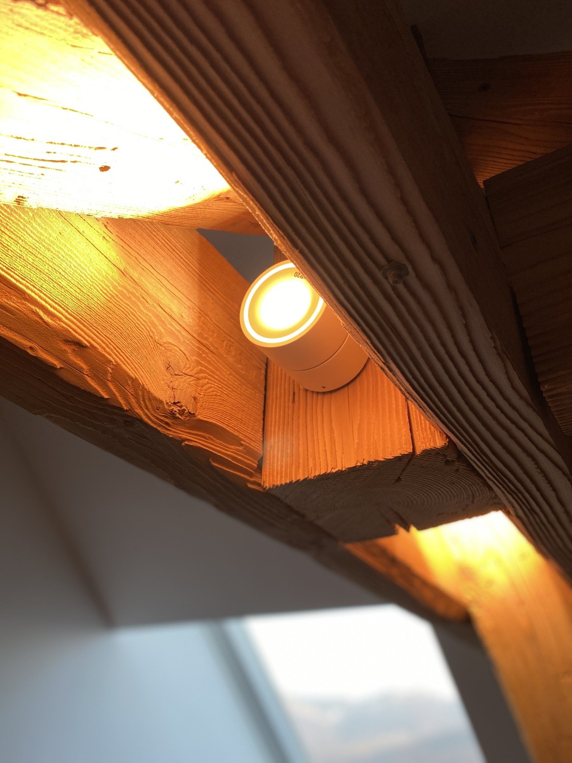 Avant-toit en bois avec deux spots encastrés allumés et la douce lumière du soleil qui filtre à travers, vue d'en bas.