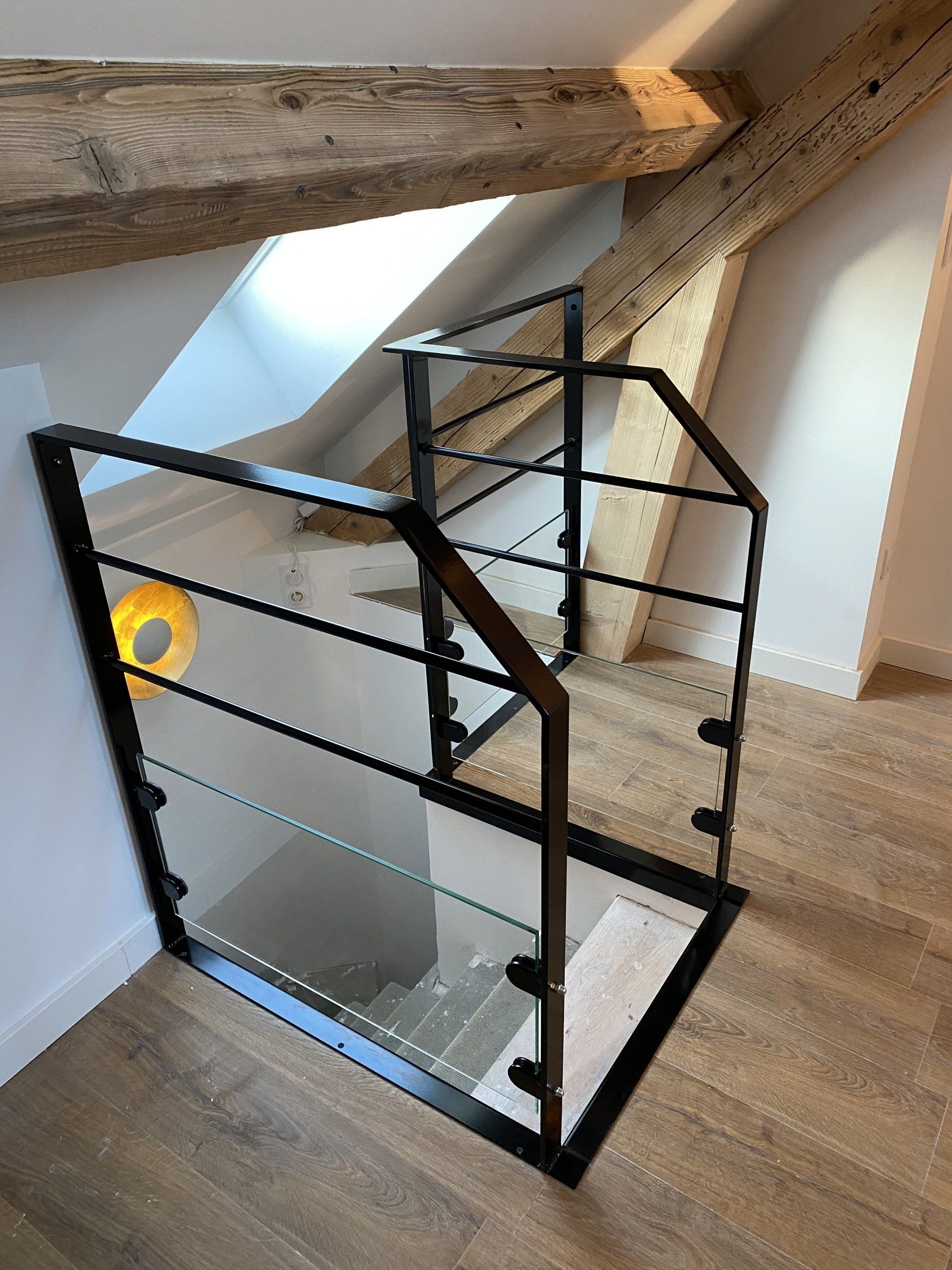 Escalier moderne menant aux combles, avec garde-corps en verre à cadre noir et poutres en bois apparentes.