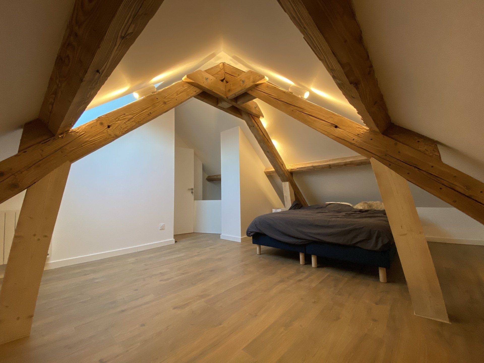 Chambre mansardée avec poutres apparentes, éclairage indirect au plafond et lit sur la droite.