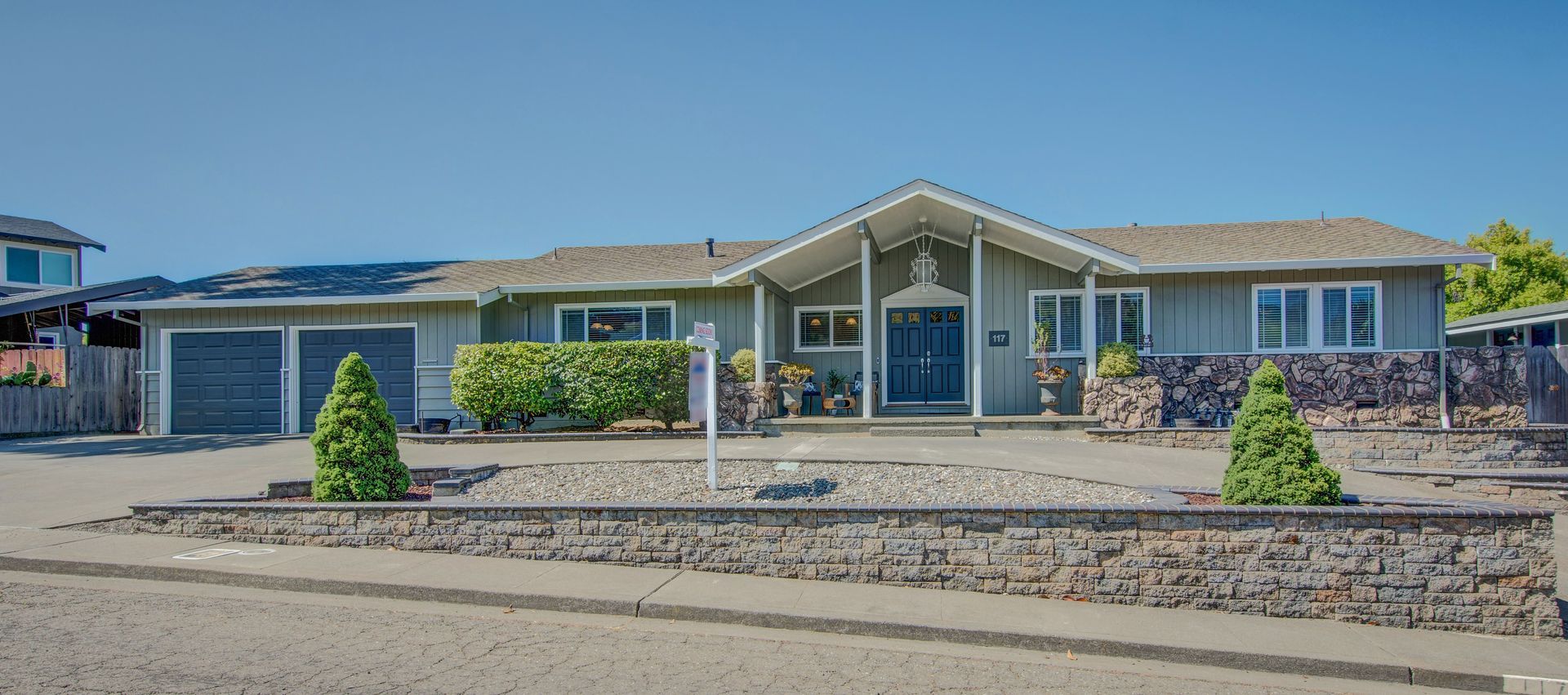 Single-story blue house with a front porch, two garage doors, and a gravel yard under a clear sky