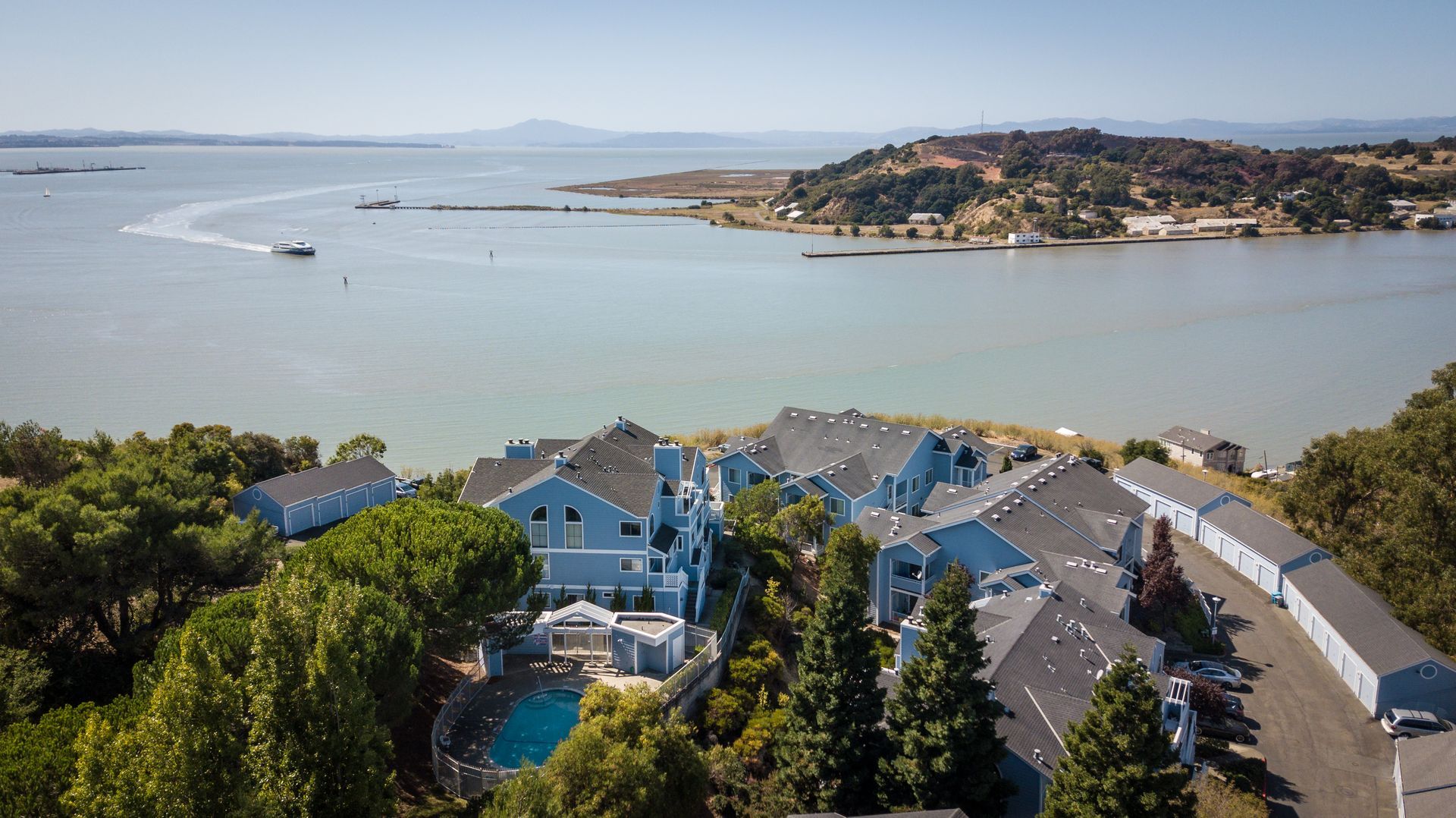 Coastal harbor town with blue-roofed houses, calm water, boats, and distant hills under a clear sky