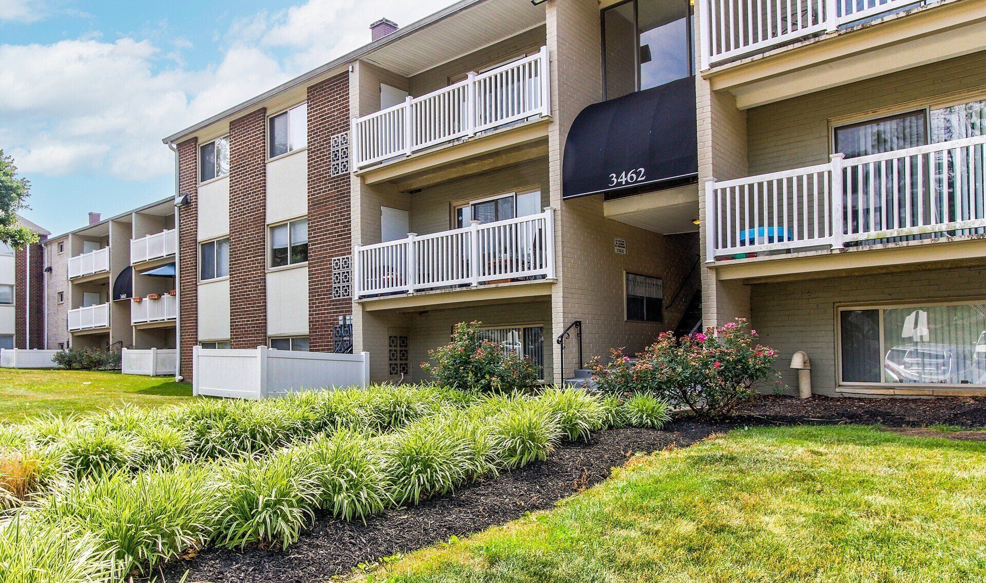 Apartment building with balconies and white railings beside a green lawn and shrubs