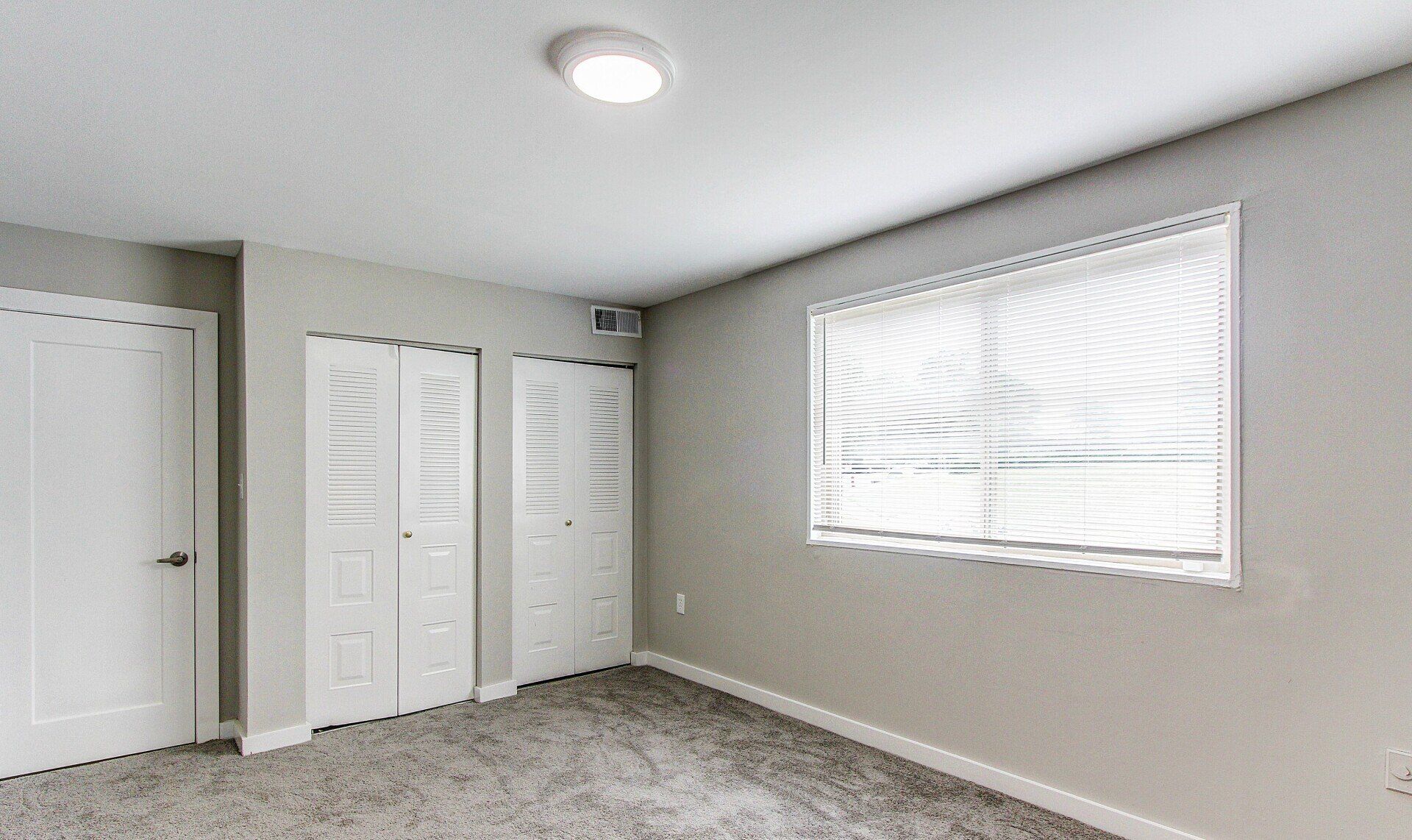 Empty beige bedroom with carpet, closet doors, and a window with blinds.