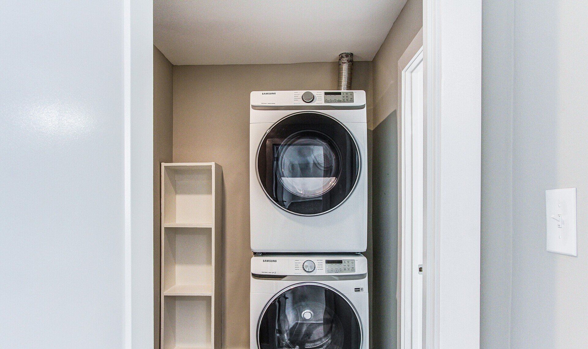 Stacked washer and dryer in a narrow laundry closet with beige walls and a small shelf