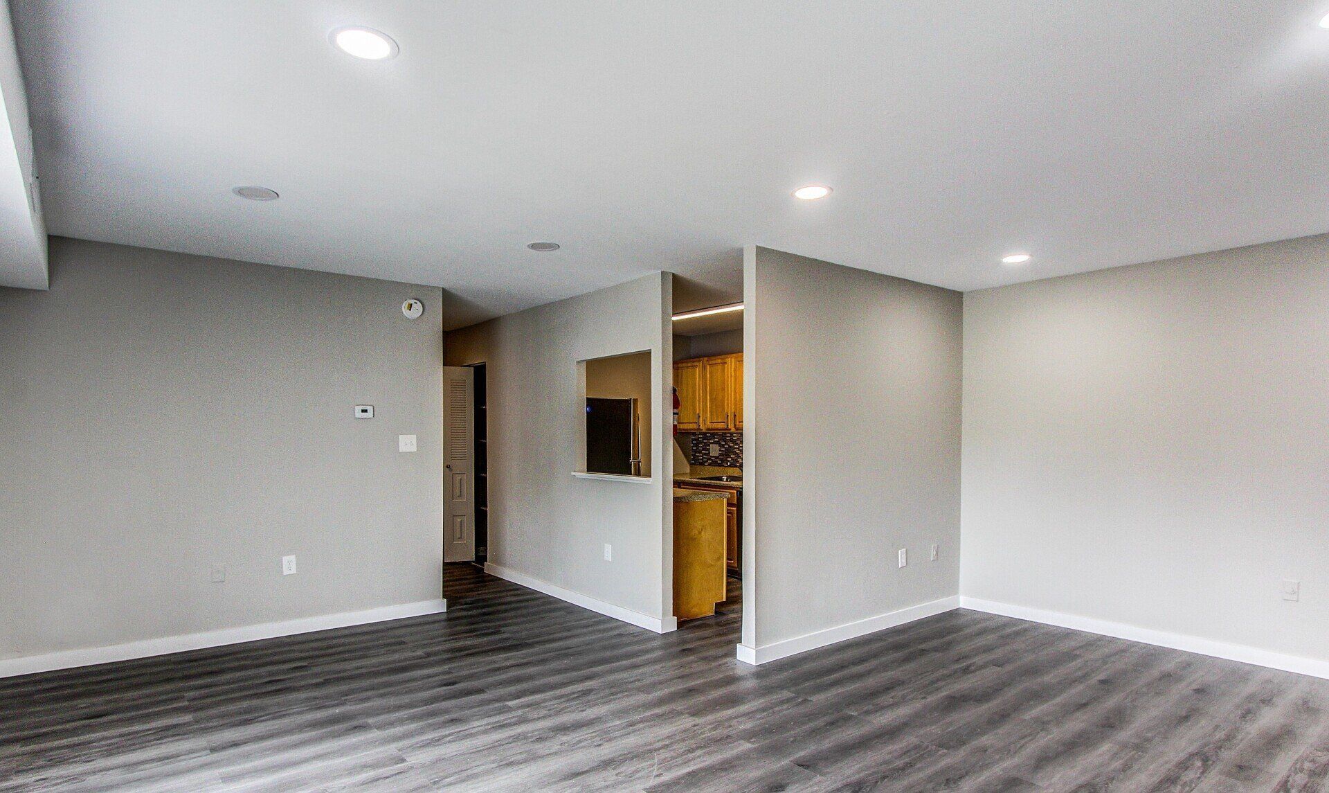 Empty modern living room with gray walls, dark wood floors, and a doorway to a kitchen area
