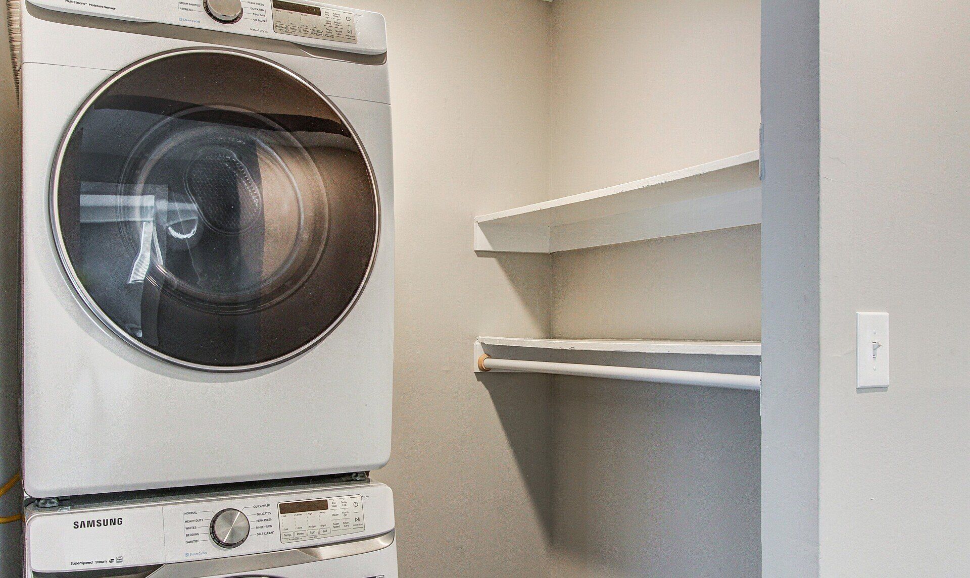 Laundry nook with stacked washer and dryer beside white wall shelves and a light switch