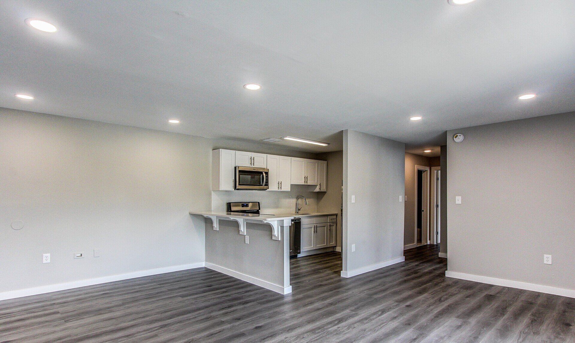 Empty modern apartment living room with open kitchen, gray wood floors, white walls, and recessed lighting