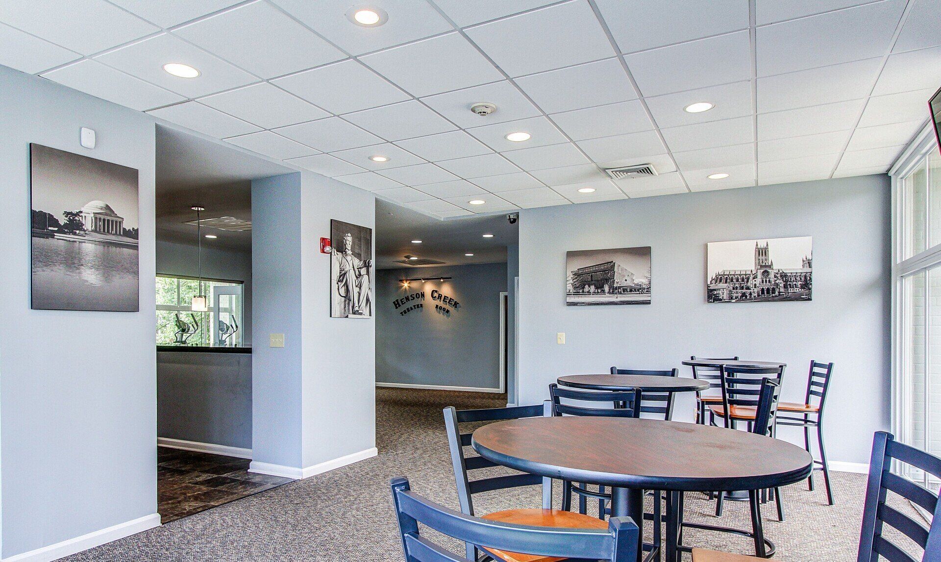 Bright office lounge with round table, chairs, gray walls, wall photos, and large windows