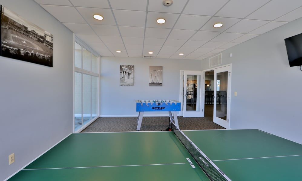 Indoor tennis court with green flooring, net, and a blue table at the far end under bright ceiling lights