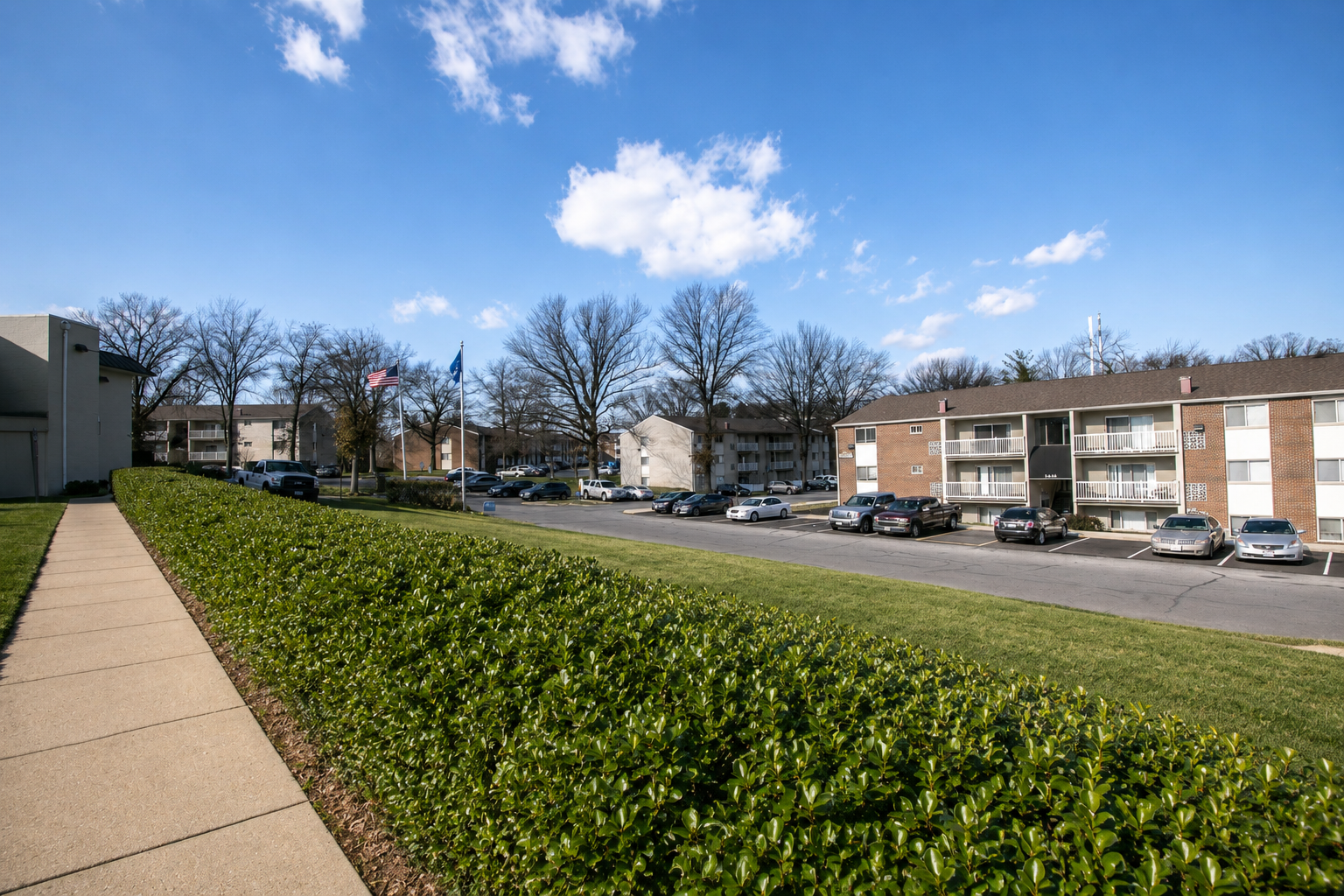 Apartment complex courtyard with sidewalk, hedge, parked cars, and blue sky