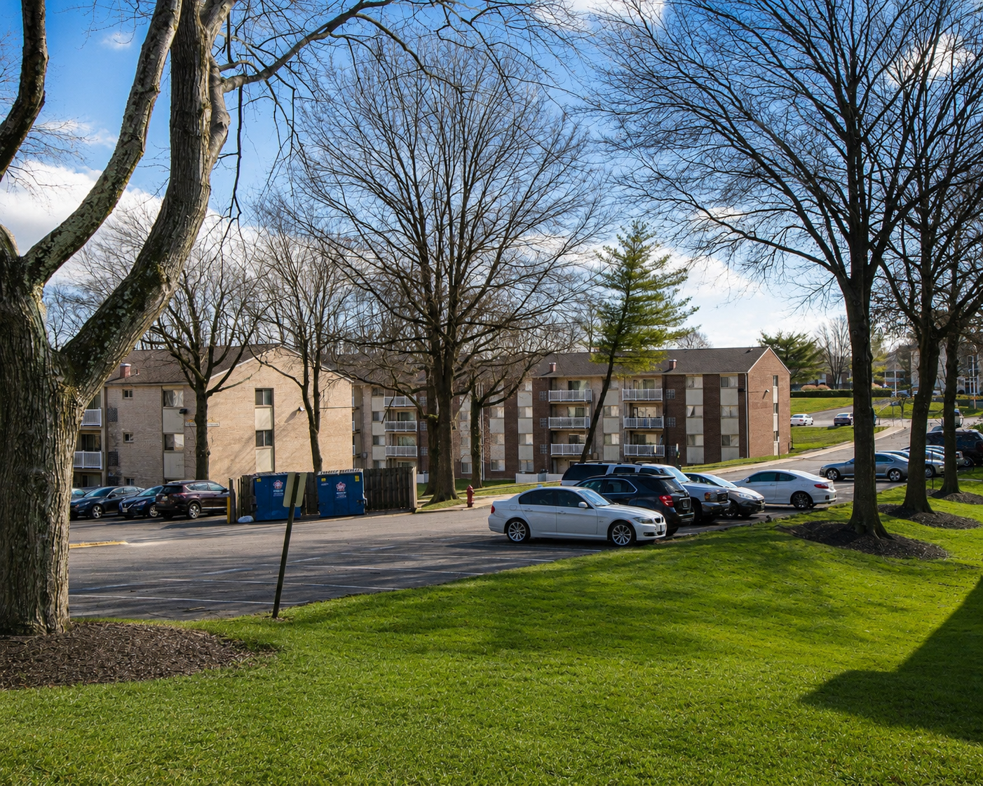 Apartment buildings behind a grassy parking lot with cars and leafless trees on a sunny day