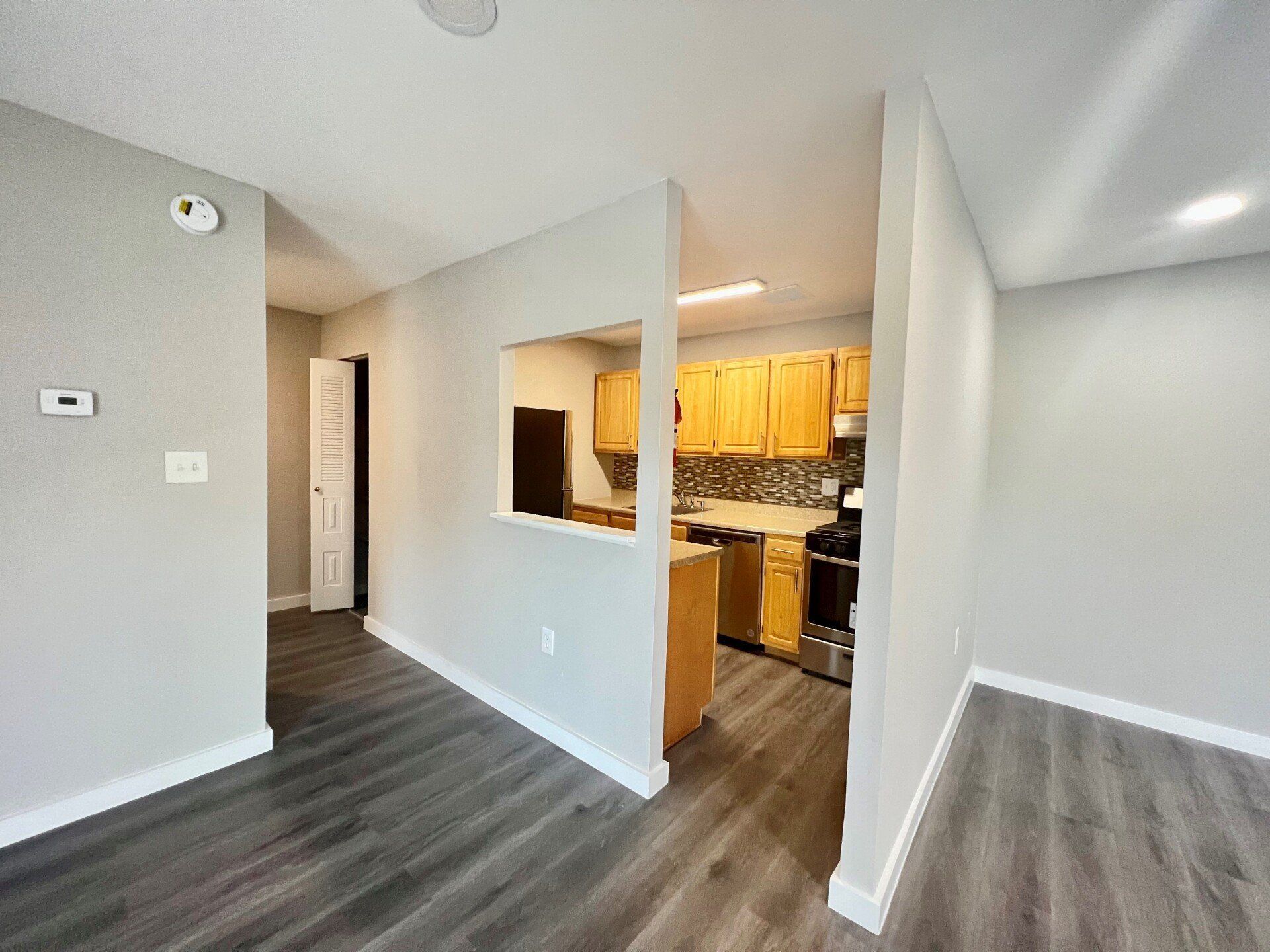 Empty apartment living area with dark flooring, white walls, and a view into the kitchen.