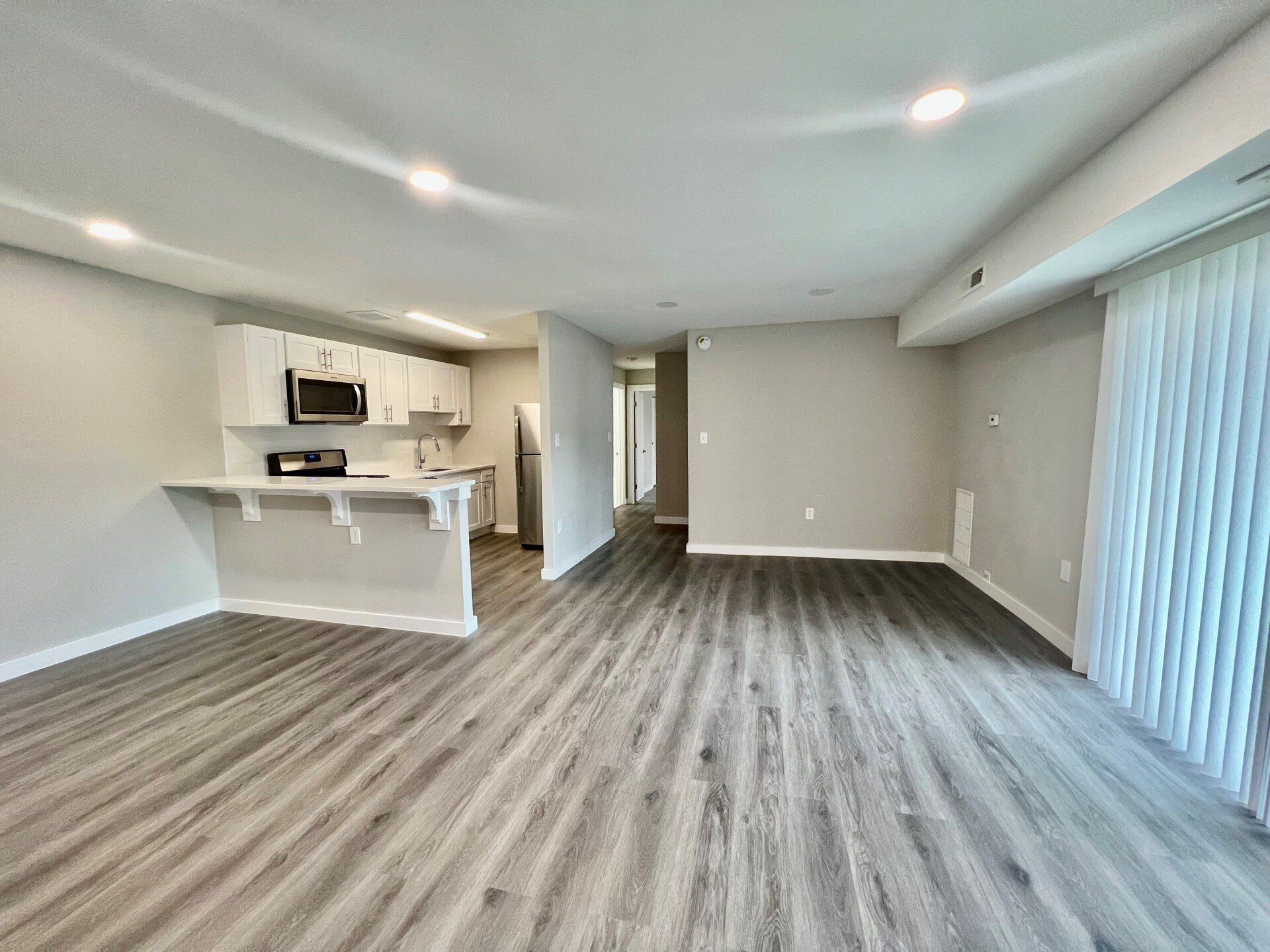 Empty modern living room with gray wood floors, white walls, and open kitchen