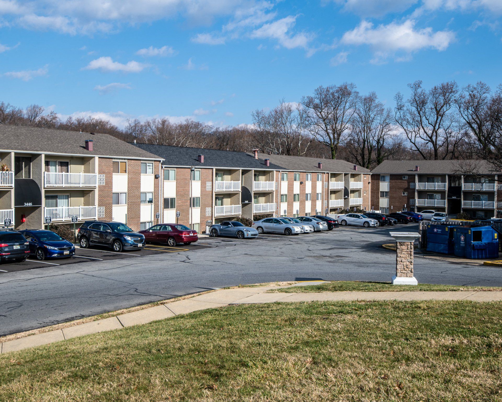 Apartment complex parking lot with parked cars, beige buildings, leafless trees, and blue recycling bins under a clear sky