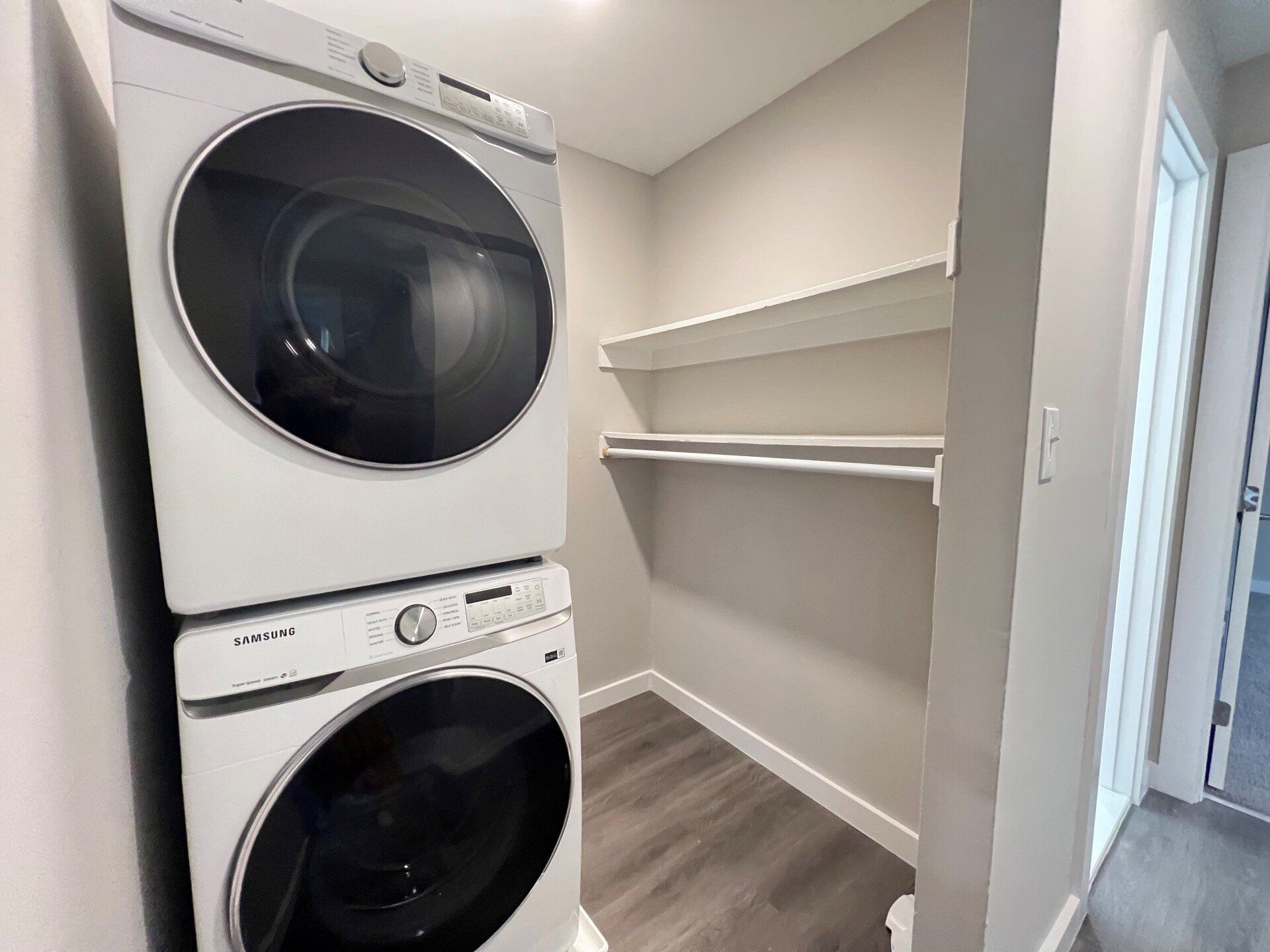 Stacked washer and dryer in a narrow laundry closet with white shelves and gray flooring.
