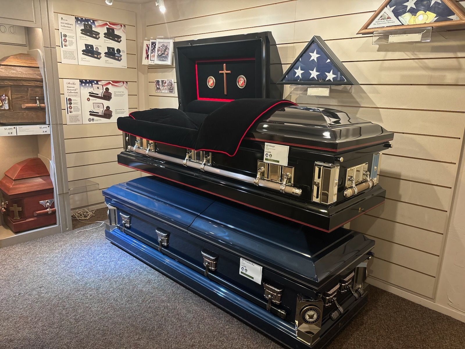 Two coffins on display in a funeral home, black and blue, silver hardware. A folded American flag is visible.