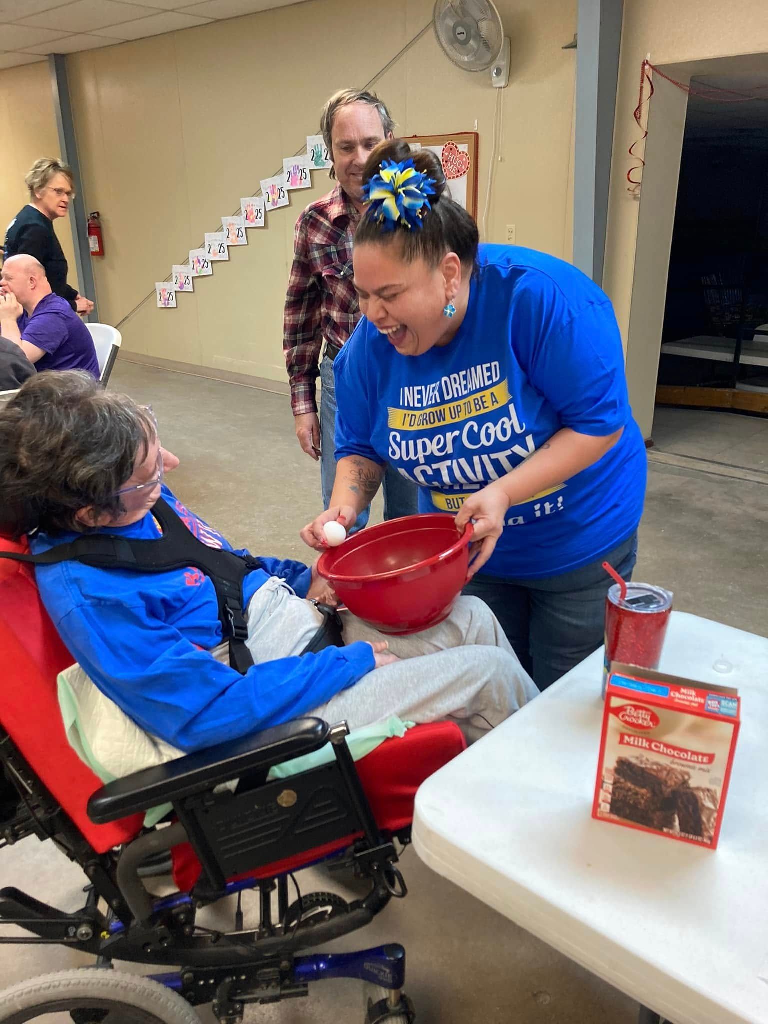 A woman in a blue shirt is holding a bowl of food next to a woman in a wheelchair.