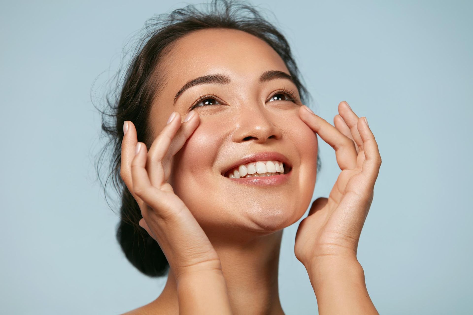 A Woman is Smiling and Touching Her Face With Her Hands — Coastal Skin & Laser in Coolum, QLD