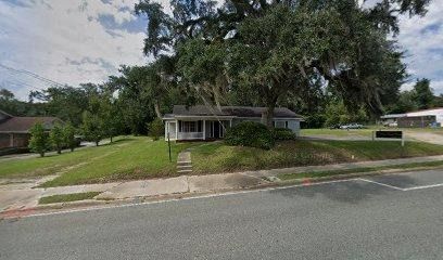 Small house on a grassy hill with a large tree. A street and a building are visible in the background.