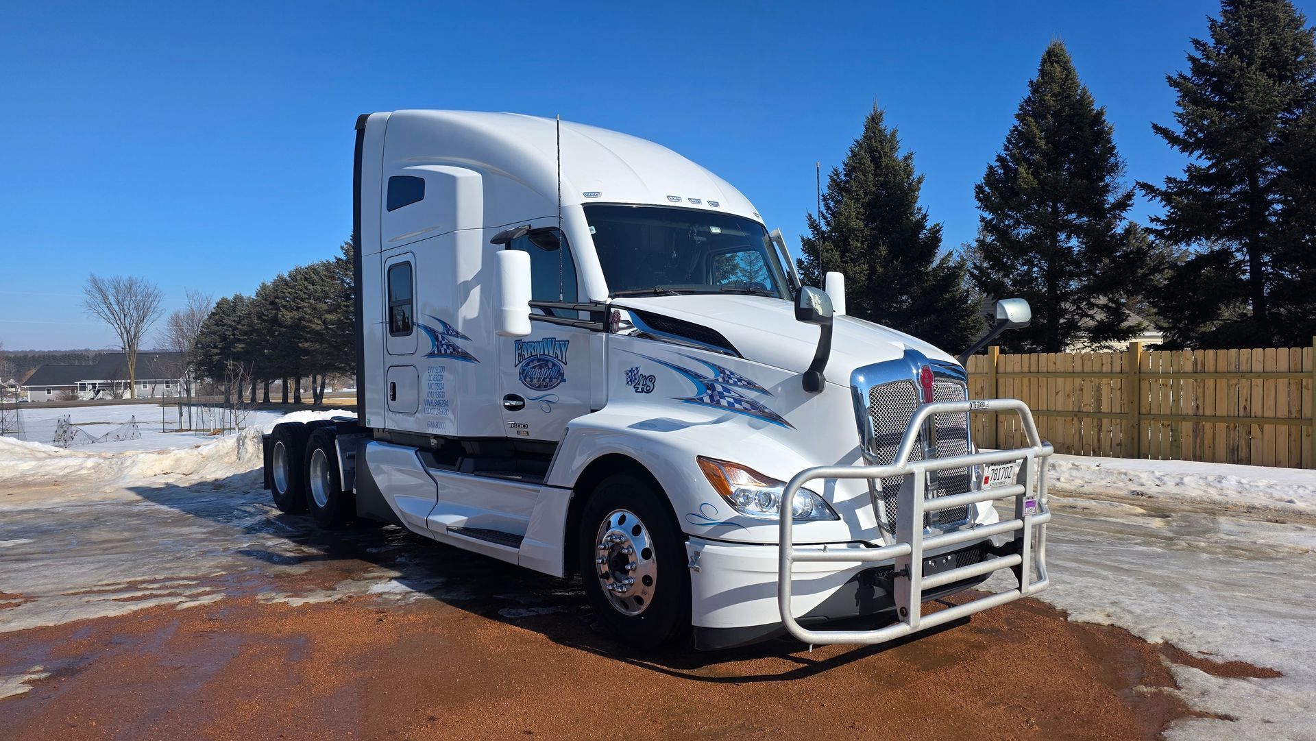 A white Kenworth semi-truck with a front-mounted grill guard parked in a snowy, sunny, outdoor lot.