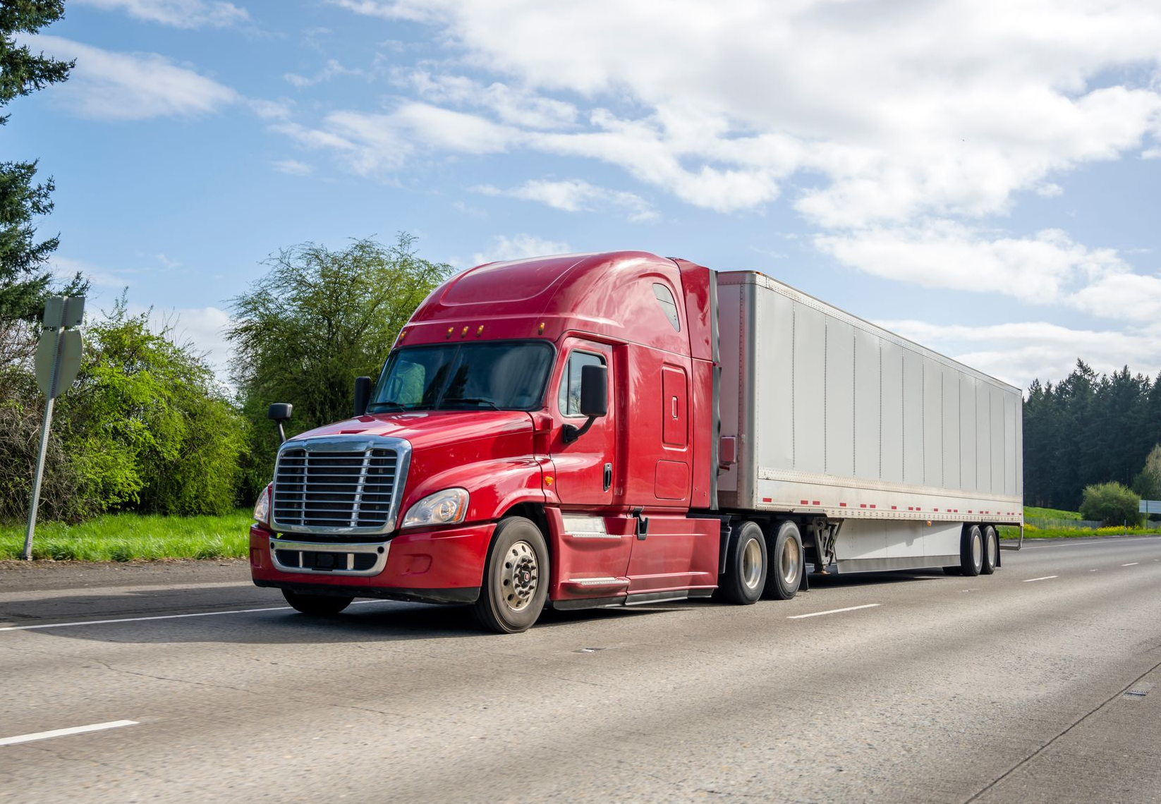 A red semi-truck with a white trailer drives down a highway on a sunny day.