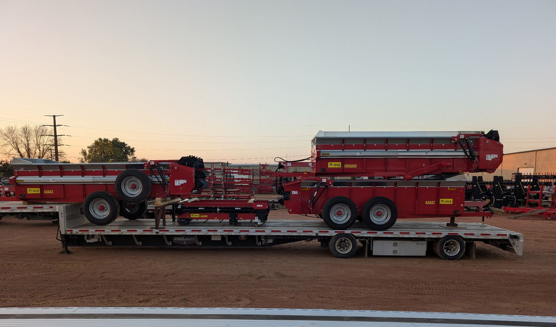 Two red agricultural trailers loaded onto a flatbed trailer at dusk.