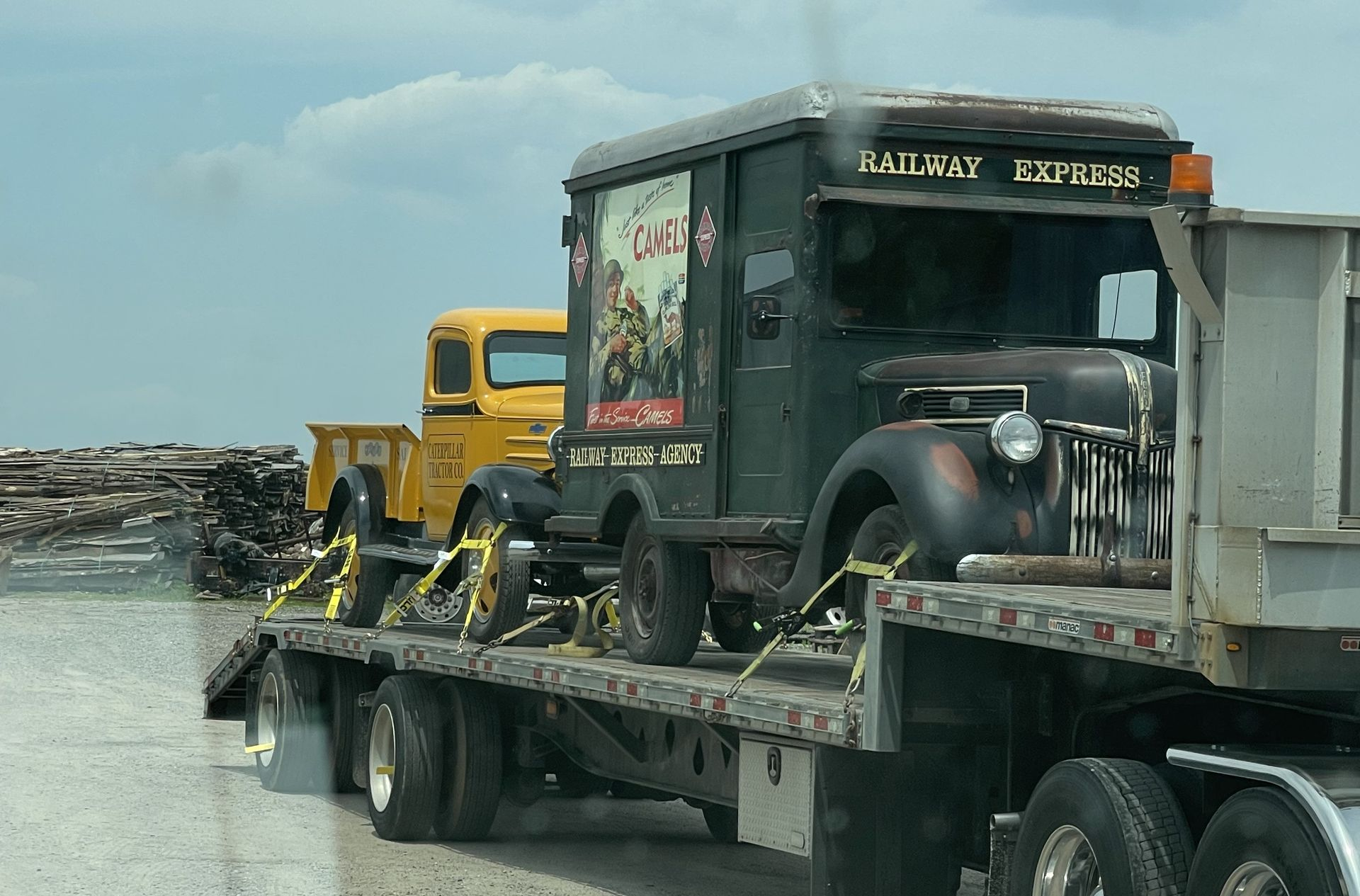 A flatbed truck carries a vintage dark green delivery truck and a yellow pickup truck along a highway.