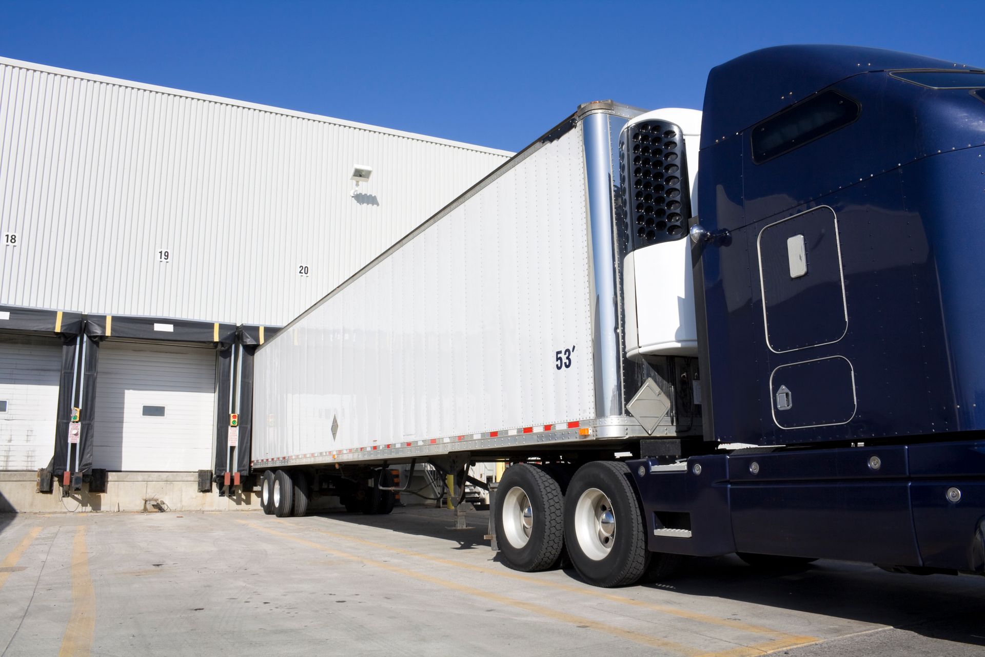 A dark blue semi-truck connected to a white refrigerated trailer parked at a loading dock against a large warehouse.