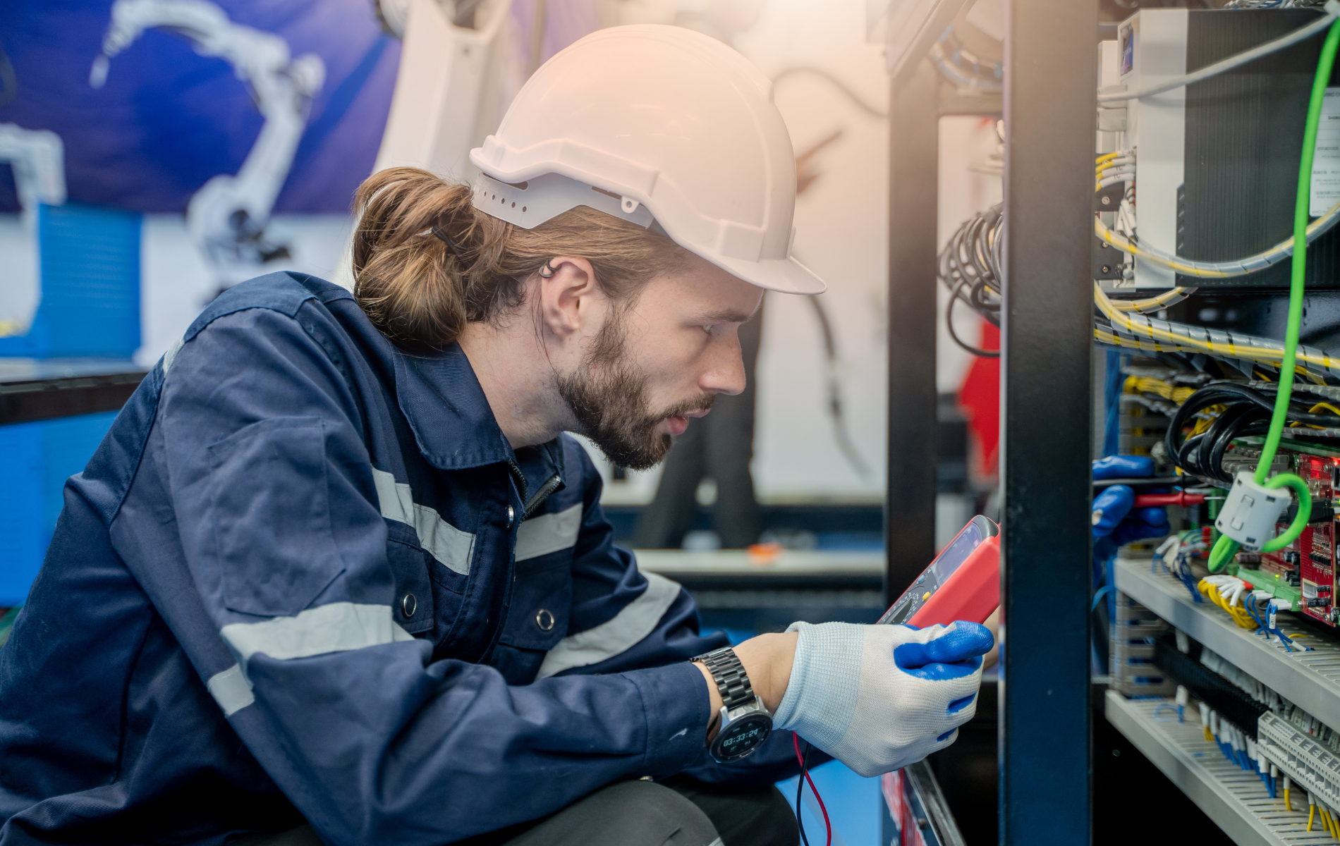 Technician in hard hat and gloves inspecting wiring inside an electrical cabinet in a workshop