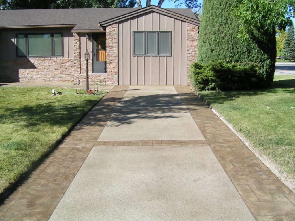 Driveway leading to a house with beige siding and brick facade. Brown stamped concrete border on either side of the driveway.