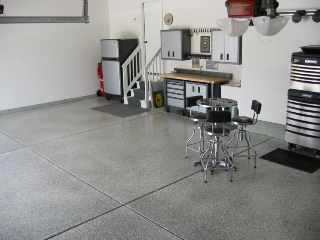 Garage interior with speckled gray floor, workbench, cabinets, refrigerator, and a small table with stools.