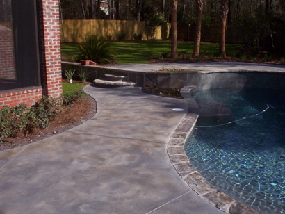 Concrete pool deck with stone coping and stairs, leading to a pool. Brick wall on the left.