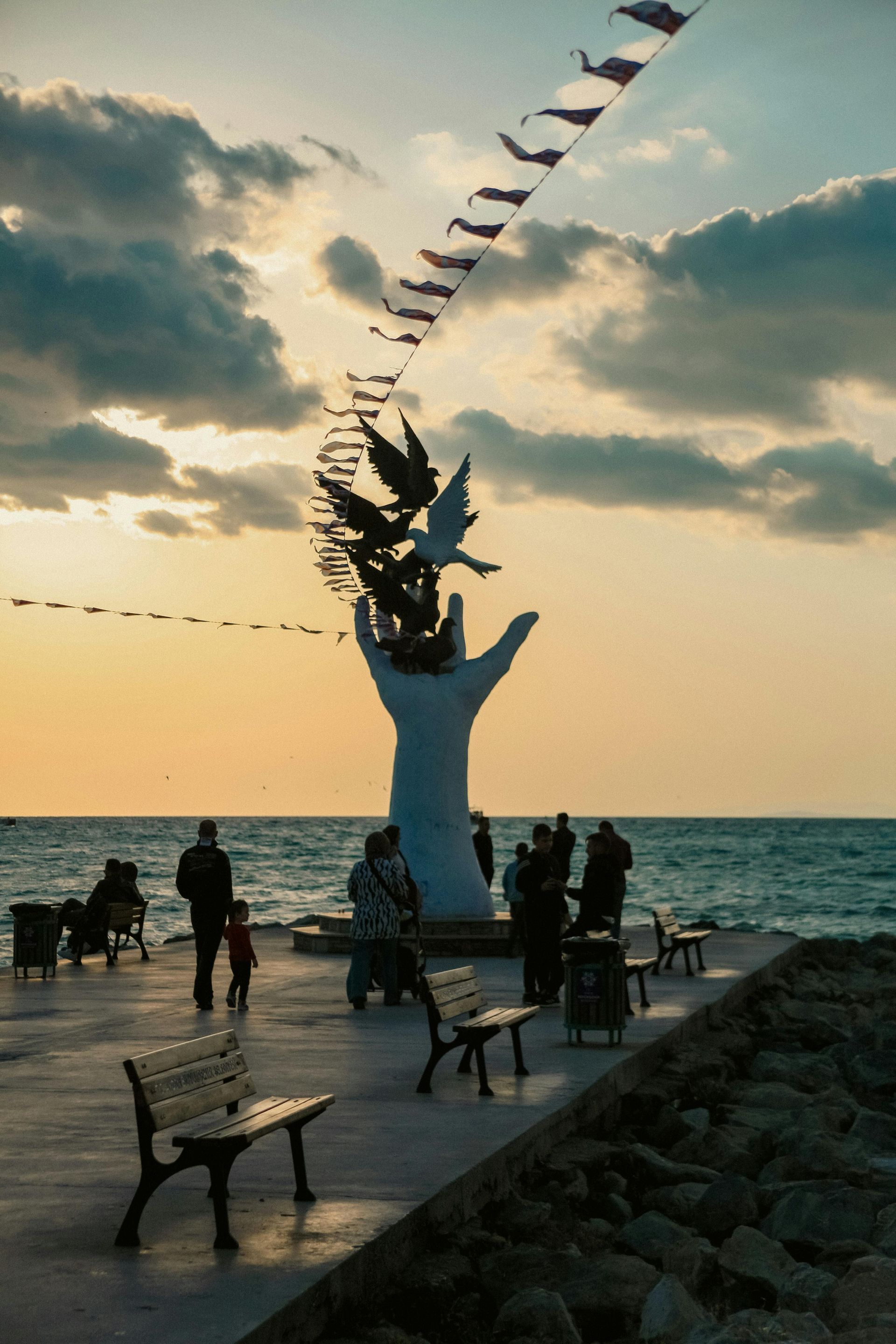 Silhouette of people on a seaside promenade at sunset beside a white statue and bunting flags