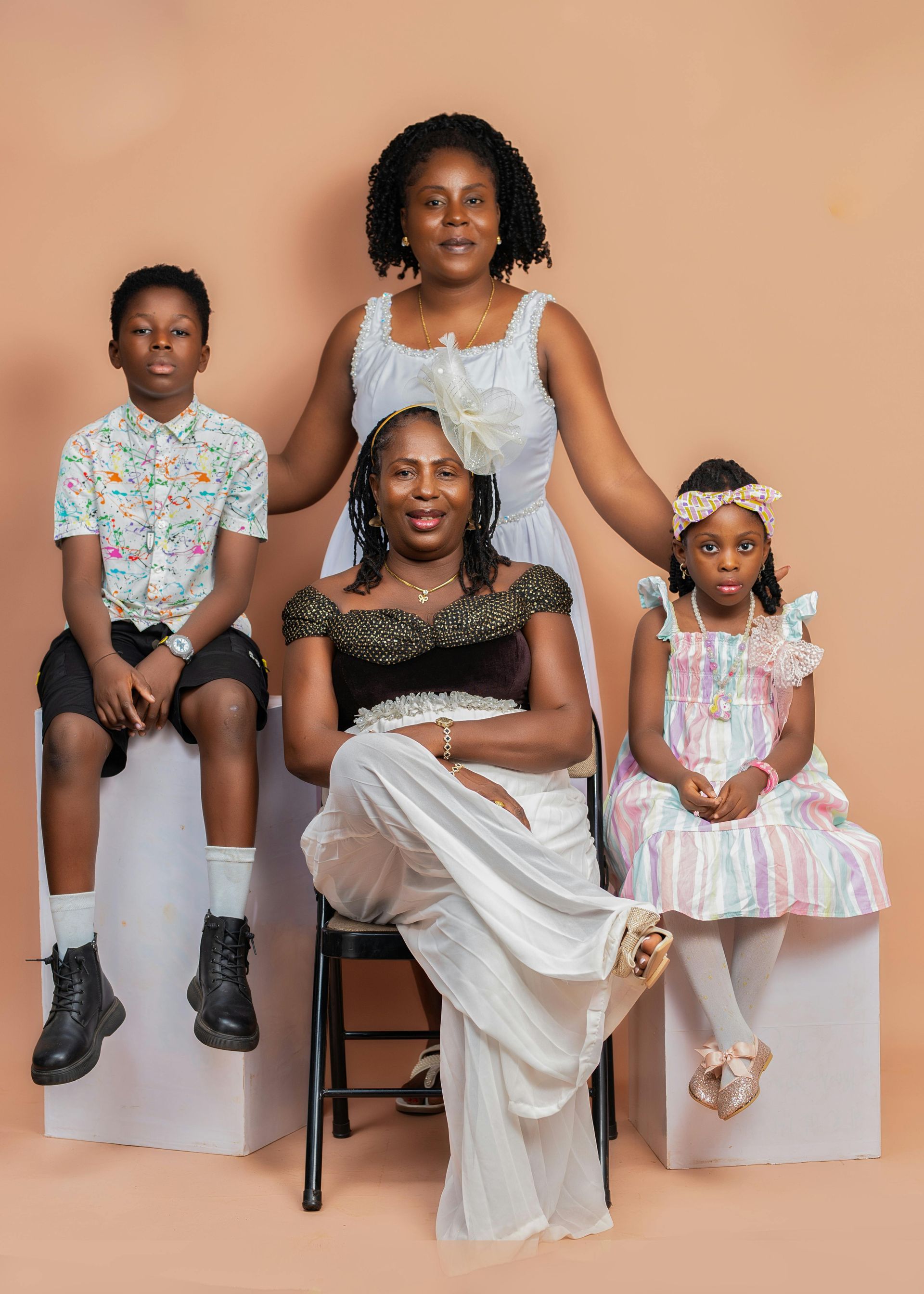 Four people posed in formal outfits against a peach backdrop, seated on white blocks with a chair centered.