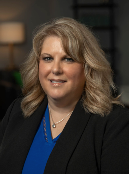 A woman in a blue shirt and black jacket is smiling for the camera.